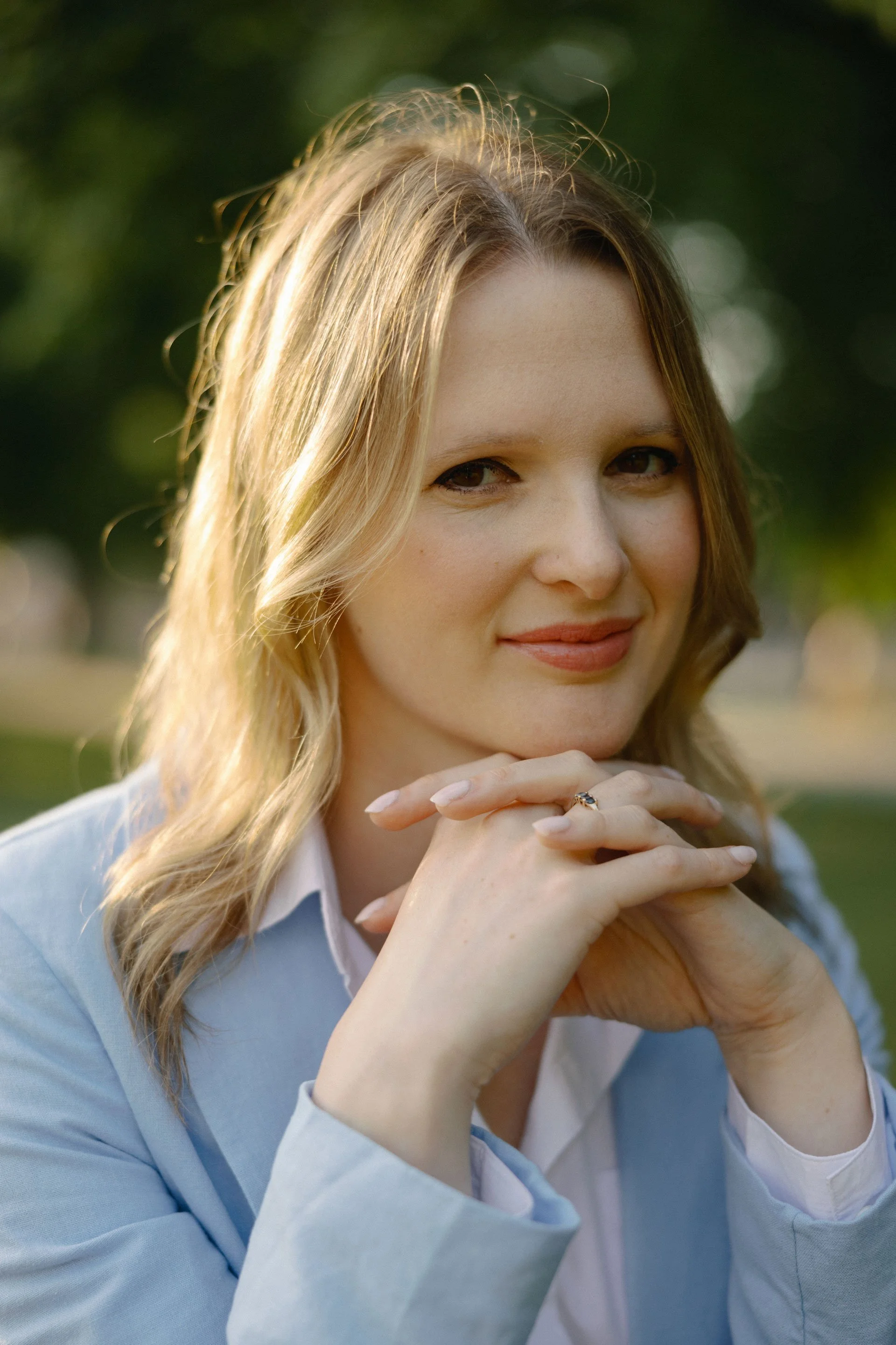 Somer Muhs, a licensed therapist, with blonde wavy hair wearing a light blue blazer and white shirt, smiling outdoors with hands clasped under her chin, greenery in the background.