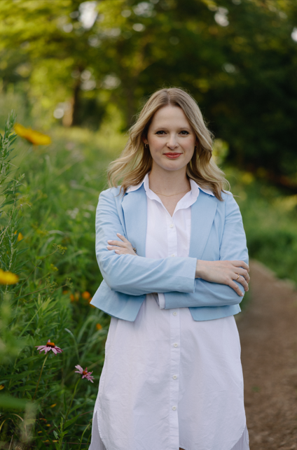 Woman with blonde hair wearing a white dress and light blue blazer standing outdoors on a dirt path with greenery and flowers in the background.