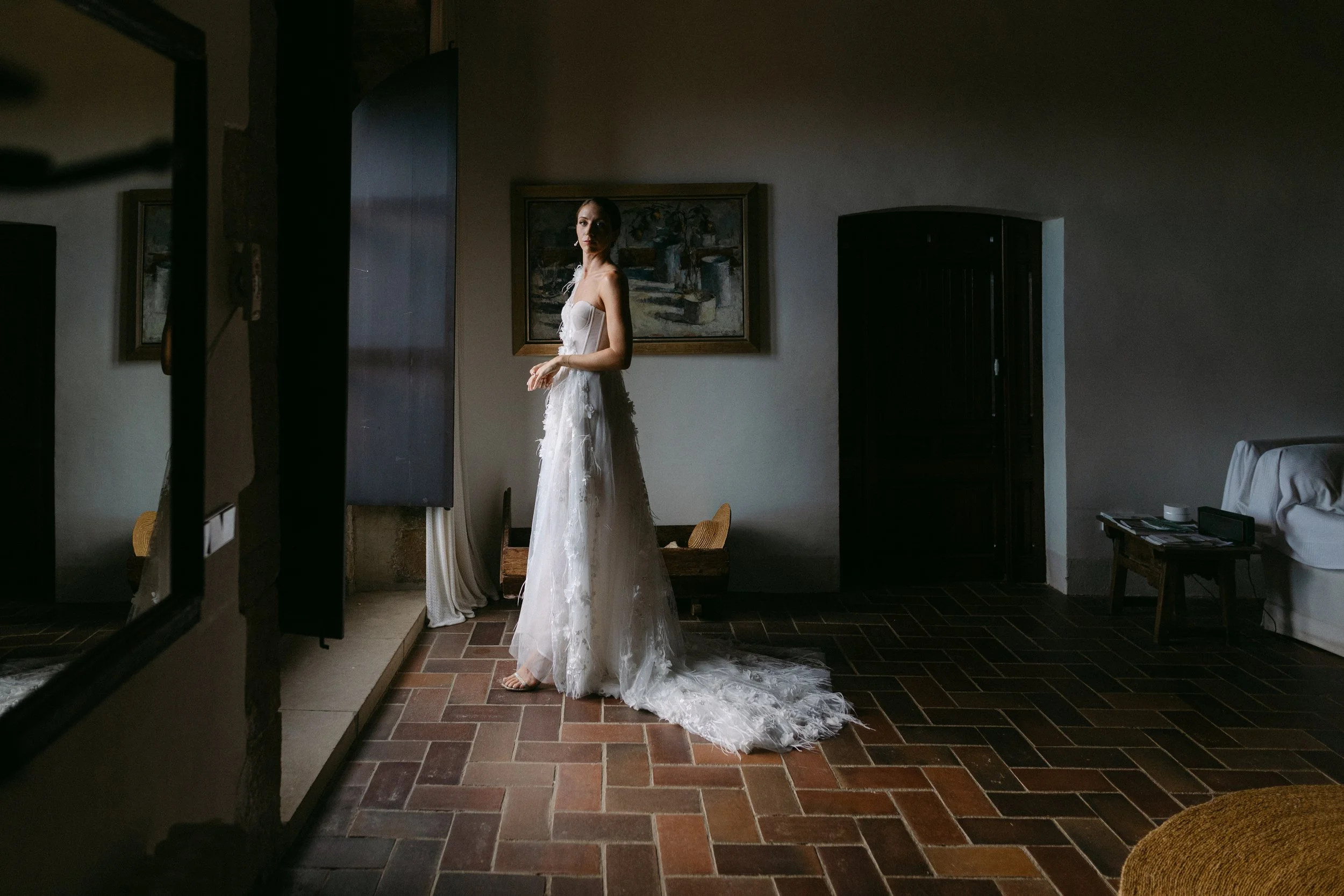 Mujer en vestido de novia de pie en interior con luz natural, en habitación con cuadros en la pared y muebles de madera.