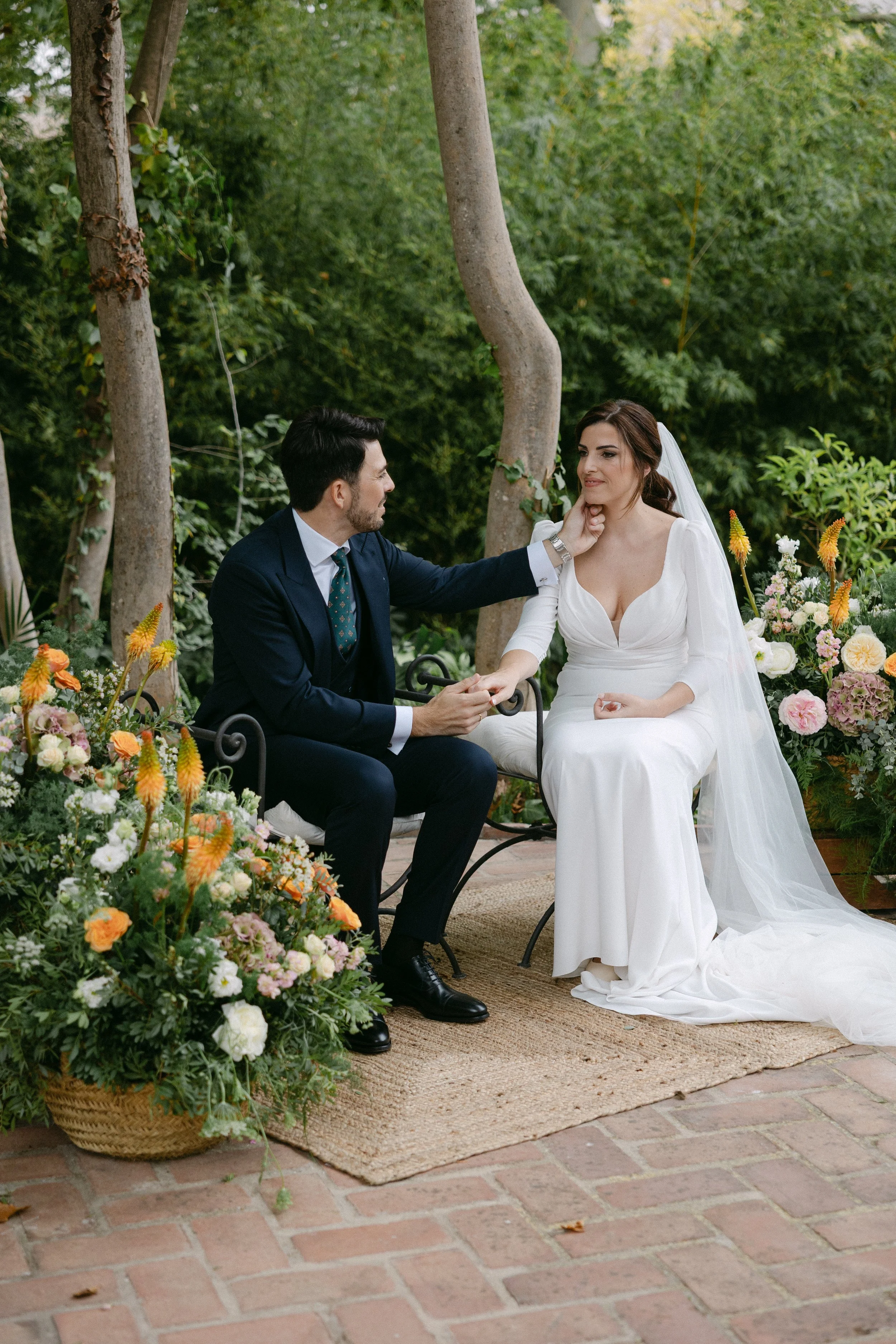 Una pareja en una boda íntima en un jardín, la mujer vestida con un vestido blanco y el hombre con un traje azul, rodeados de flores y árboles.
