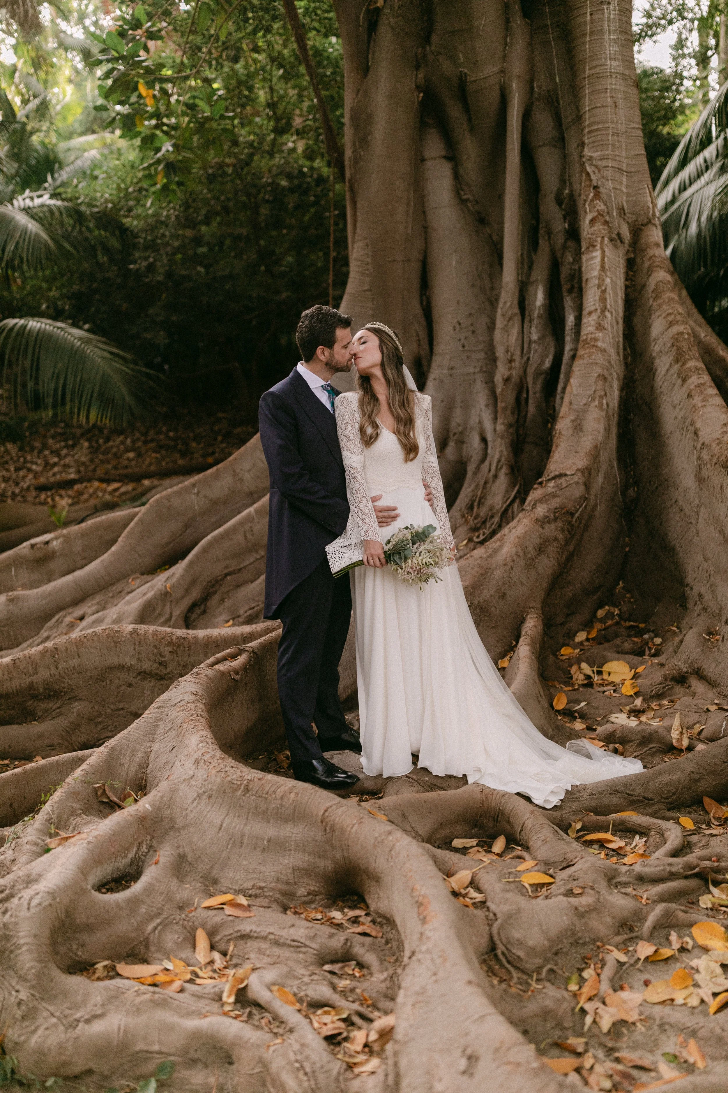Pareja de novios en un bosque, la novia con vestido blanco y ramo, el novio con traje oscuro, tocando frente a un árbol grande con raíces expuestas.
