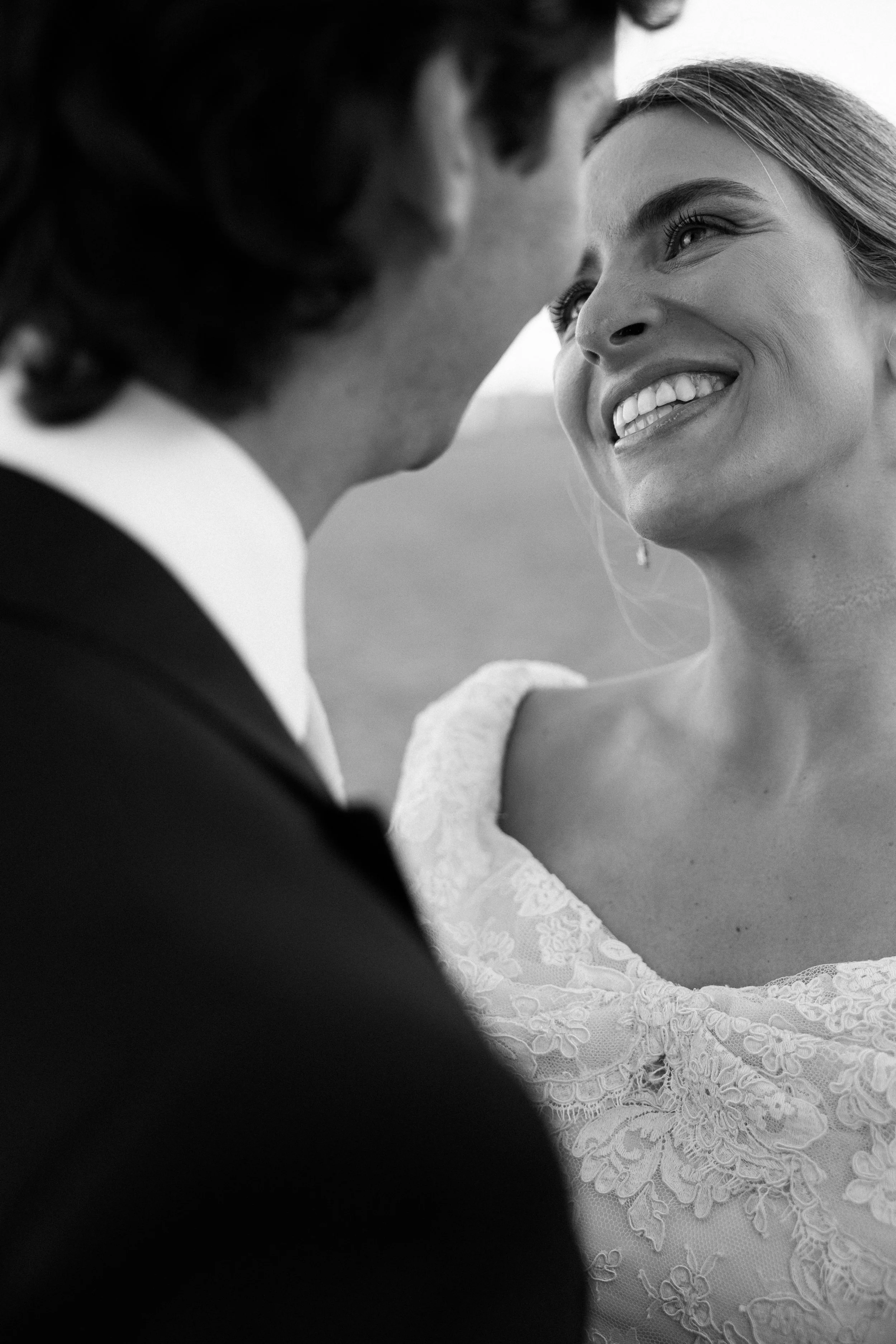 Una pareja de bodas en un momento íntimo, la mujer sonriendo y la vista parcial del rostro del hombre en un entorno al aire libre.