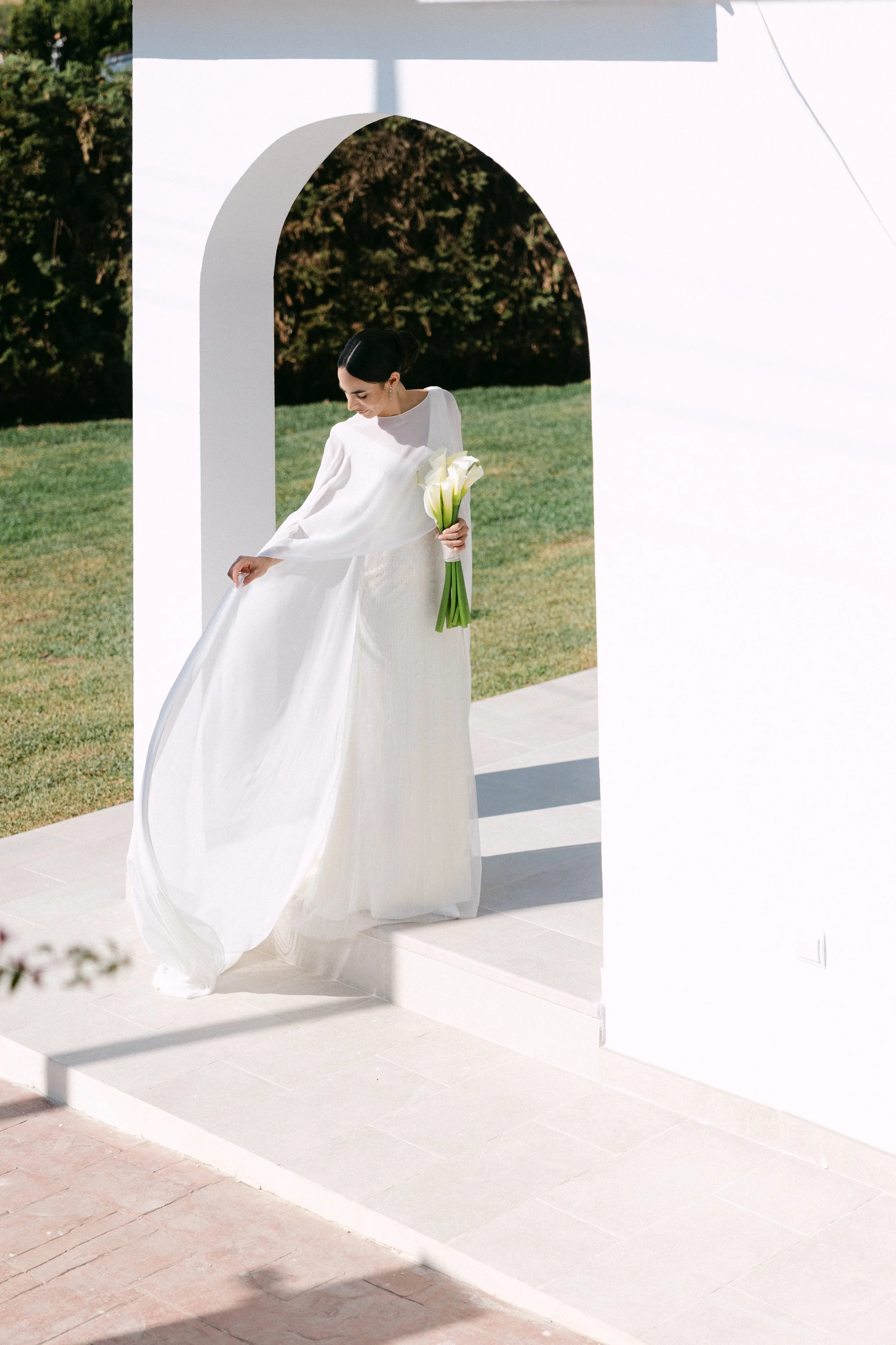 Mujer vestida con un vestido de novia blanco, sosteniendo un ramo de flores blancas, en un escenario exterior con estructura blanca y césped verde.