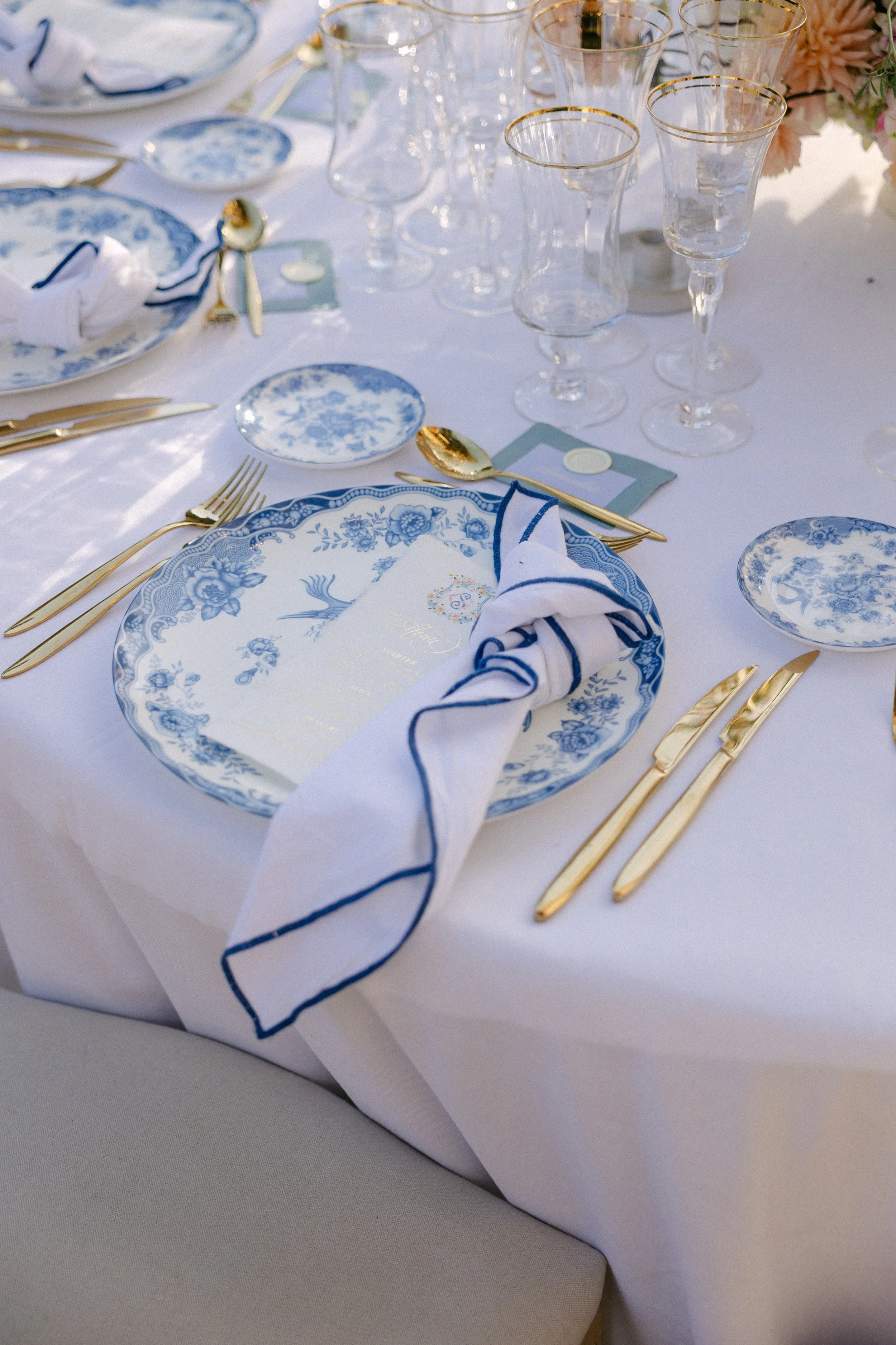 Mesa elegant decorada con platos de cerámica azul y blanco, cubiertos dorados, copas de vino con borde dorado, y un servilleta de tela blanca con borde azul, organizada para una celebración sofisticada.