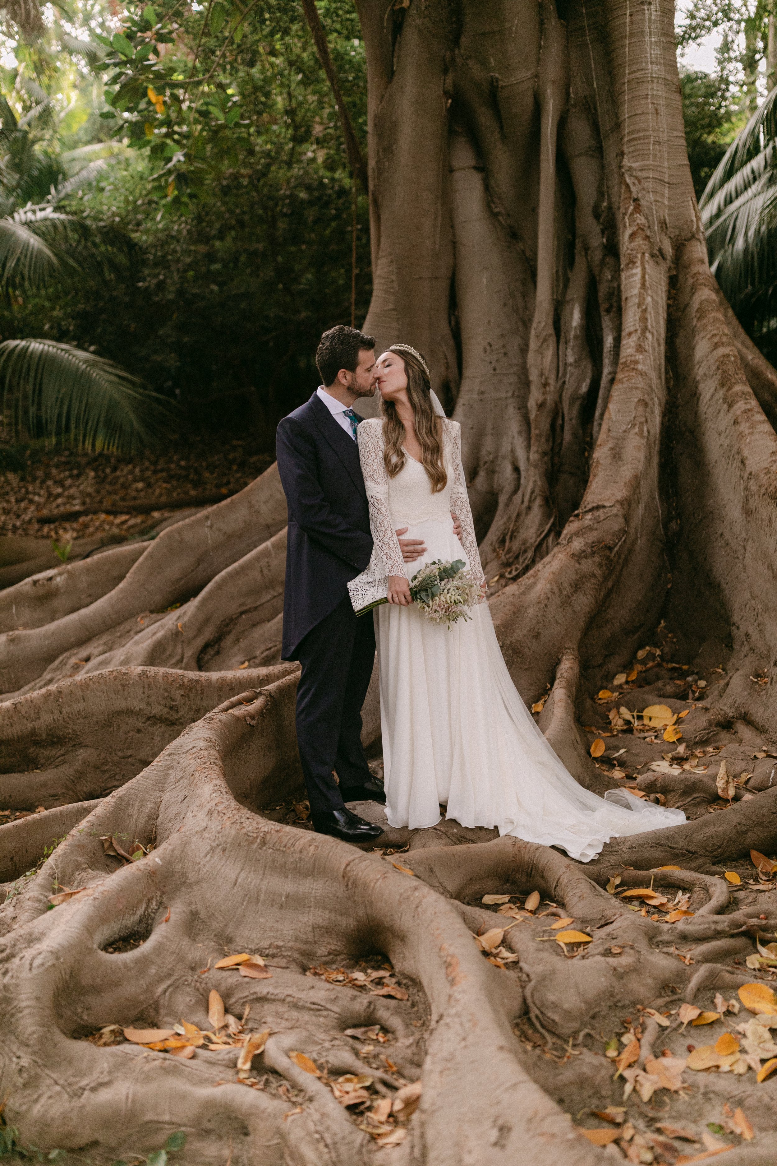 Pareja de novios en un bosque, con raíces grandes y un árbol enorme de fondo, la novia lleva vestido blanco y ramo, el novio traje oscuro.