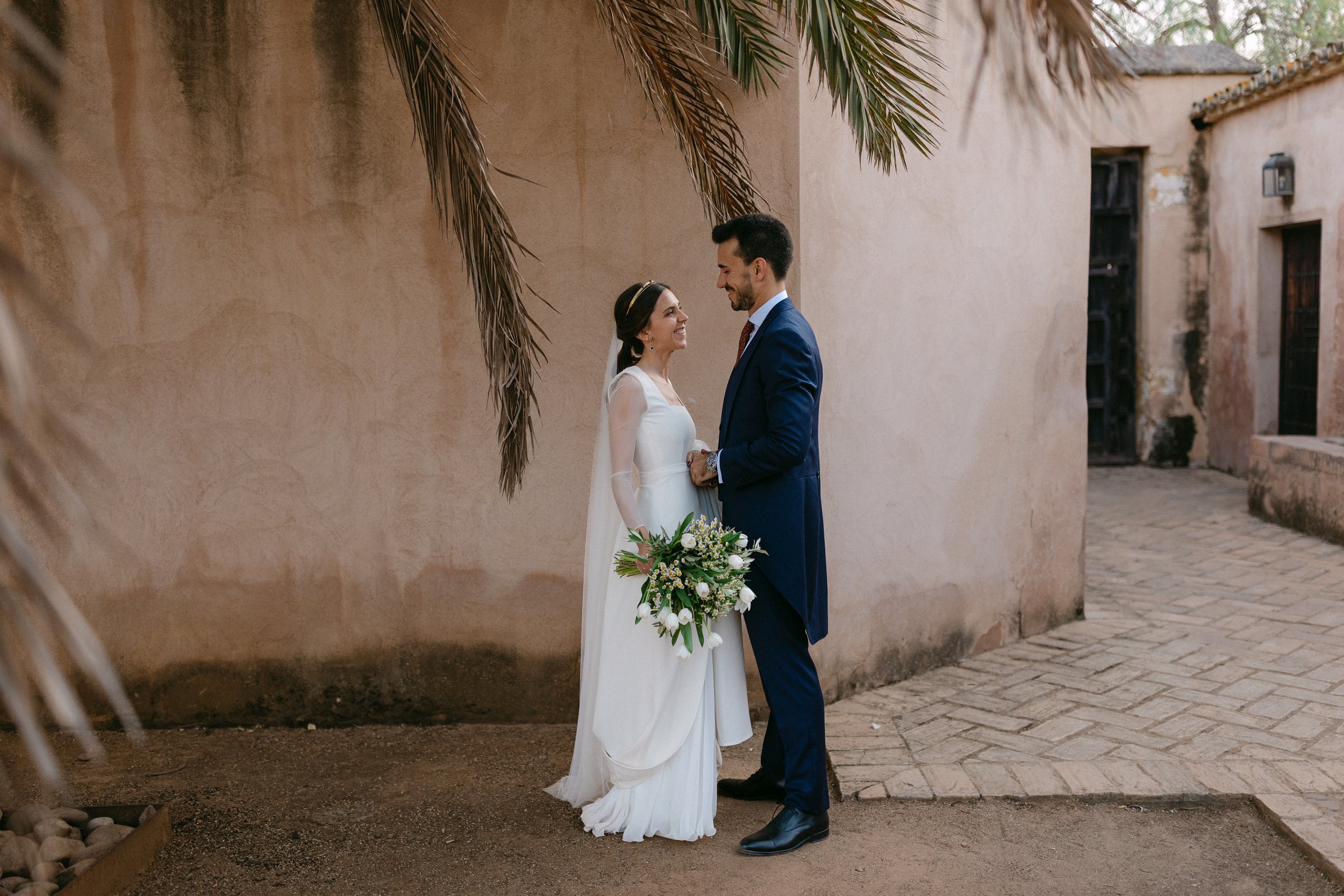 Pareja de recién casados en portada, la mujer en vestido blanco con ramo de flores y el hombre en traje azul, de pie frente a una pared de color beige, en un patio con suelo de piedra.