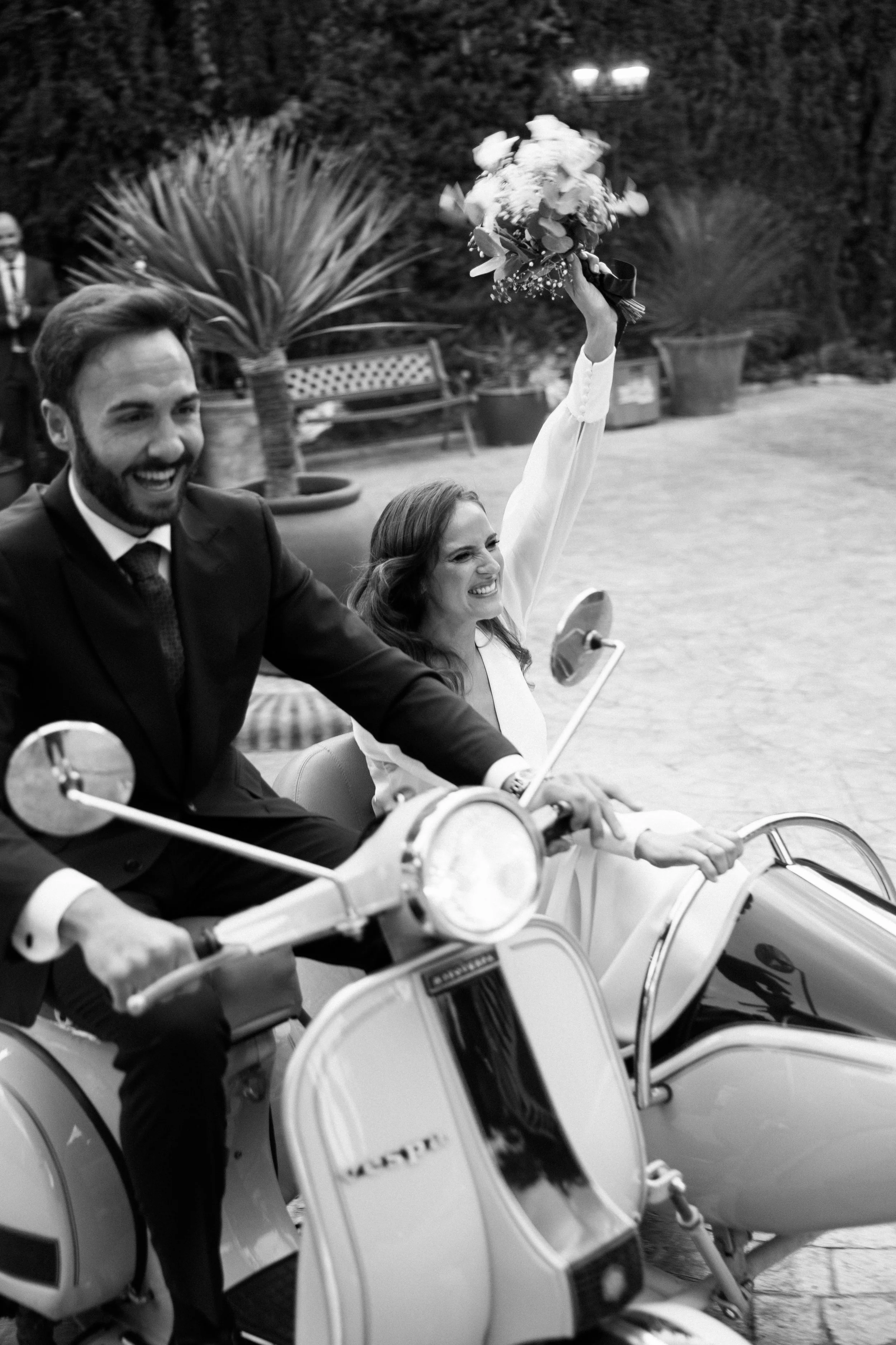 Una pareja feliz en una motocicleta con sidecar, ella sostiene un ramo de flores y está sonriendo, en un entorno al aire libre con plantas y bancas.