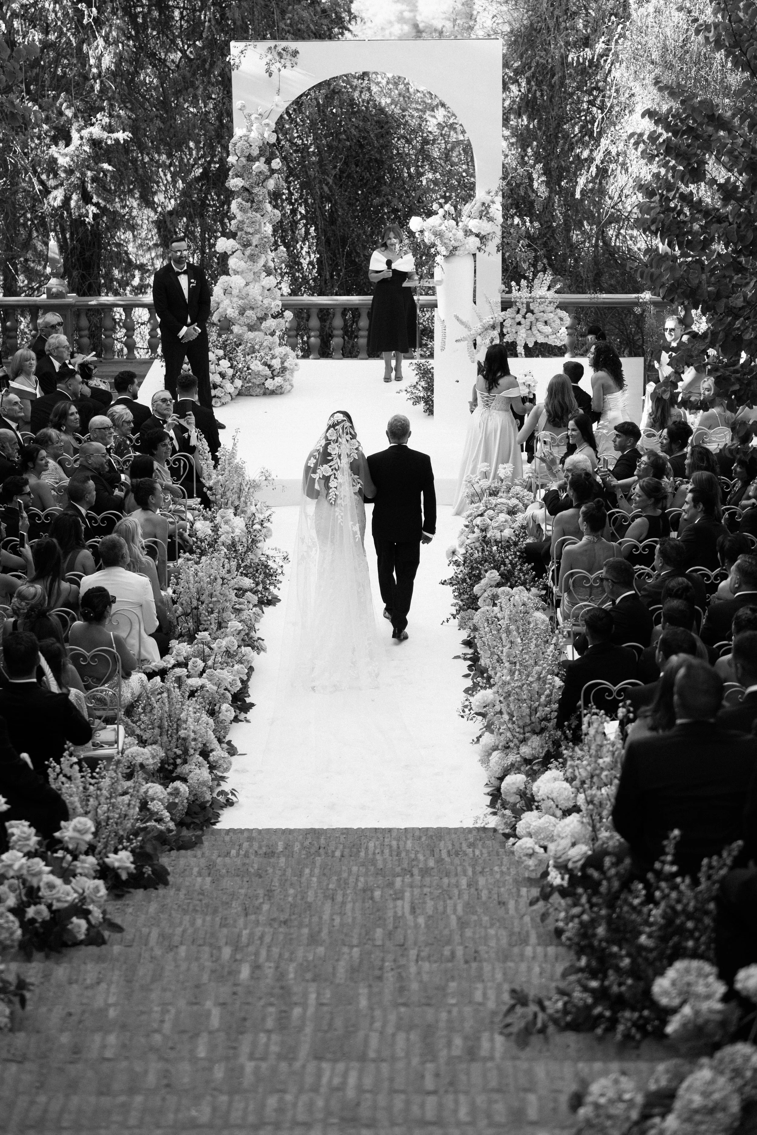 Boda al aire libre con pareja de recién casados caminando por el pasillo, rodeados de invitados y decoraciones florales.