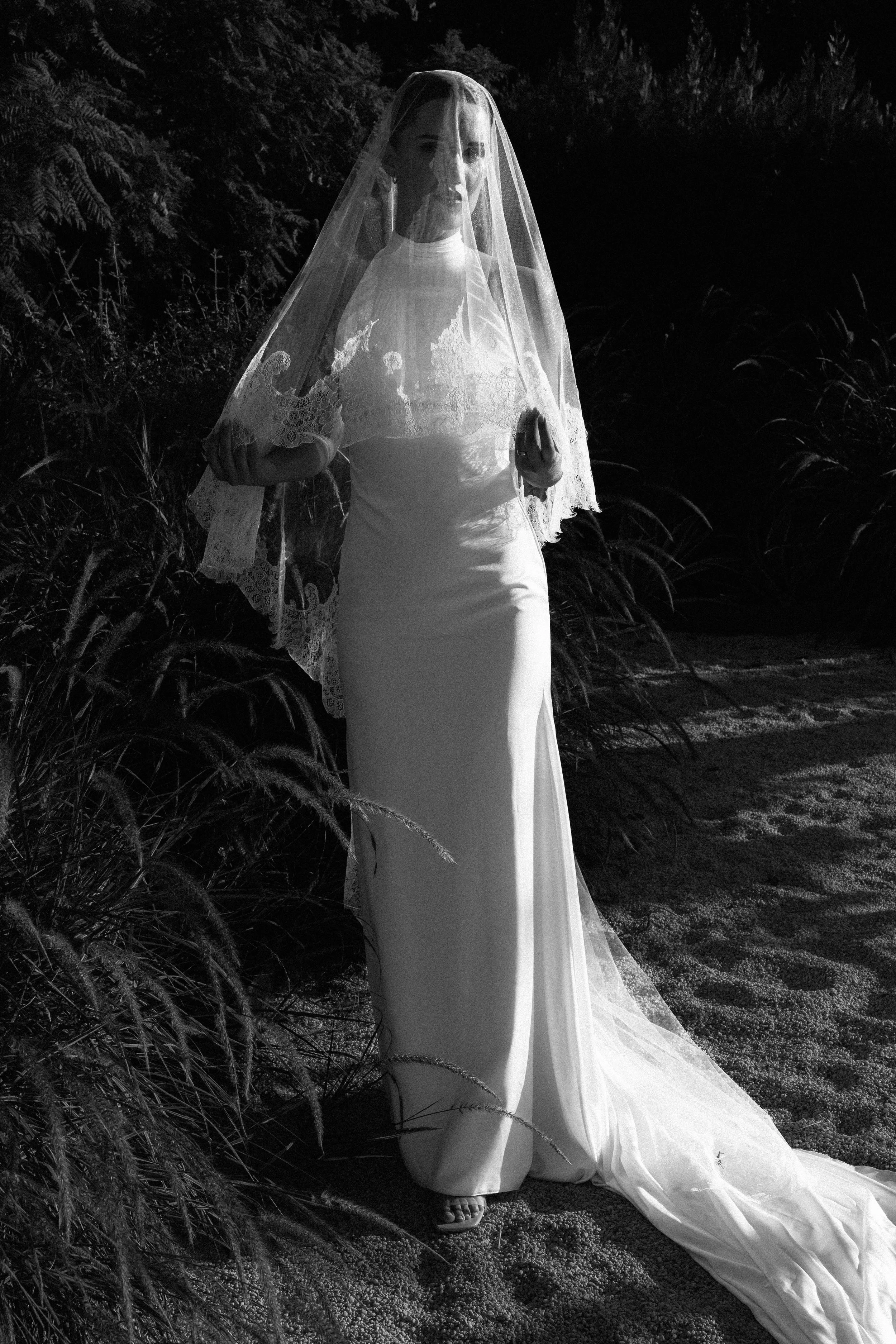 Mujer vestida de novia con velo, de pie en un entorno natural, en blanco y negro.