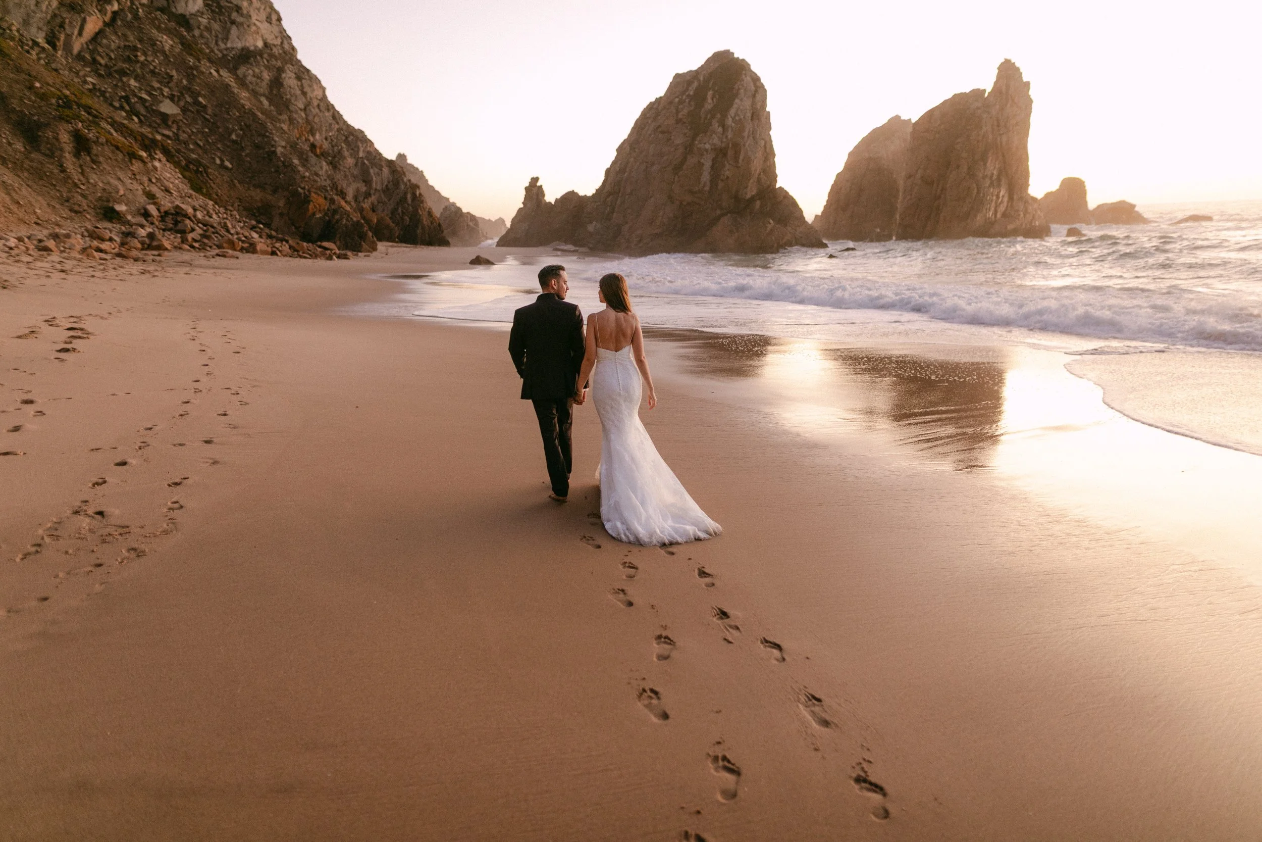 Pareja de novios caminando por la playa con rocas y el mar al atardecer.