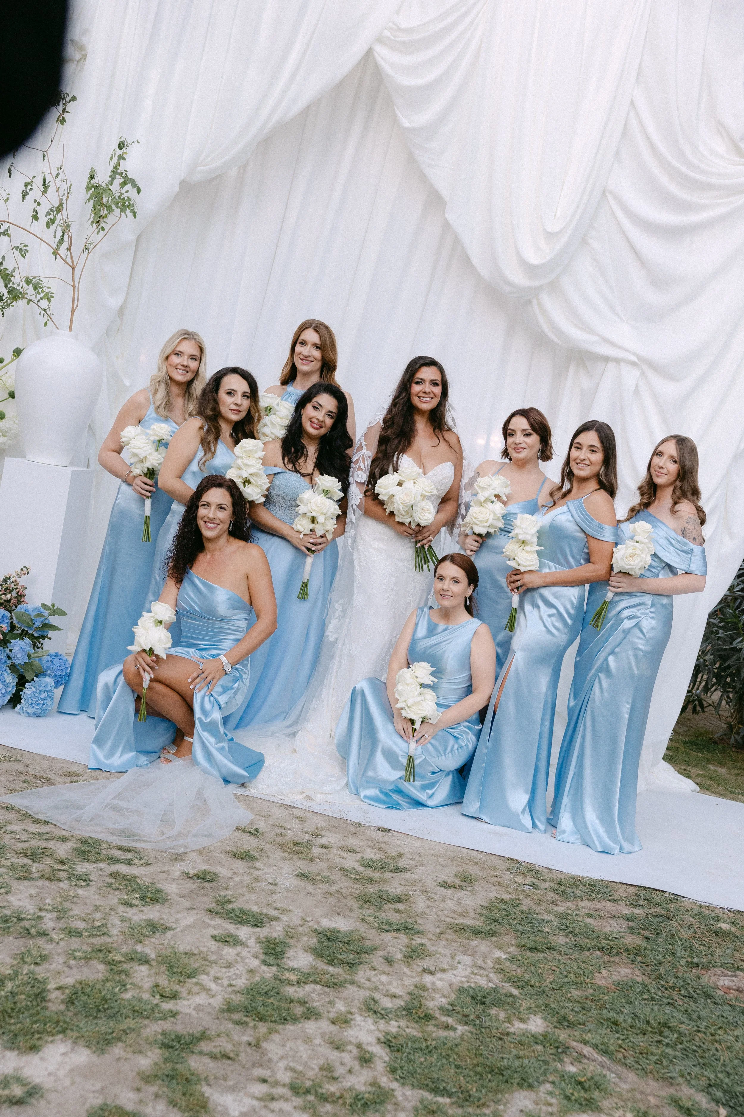 Grupo de mujeres en vestido de novia y damas de honor posando en una ceremonia de boda al aire libre decorada con cortinas blancas y flores blancas y azules.