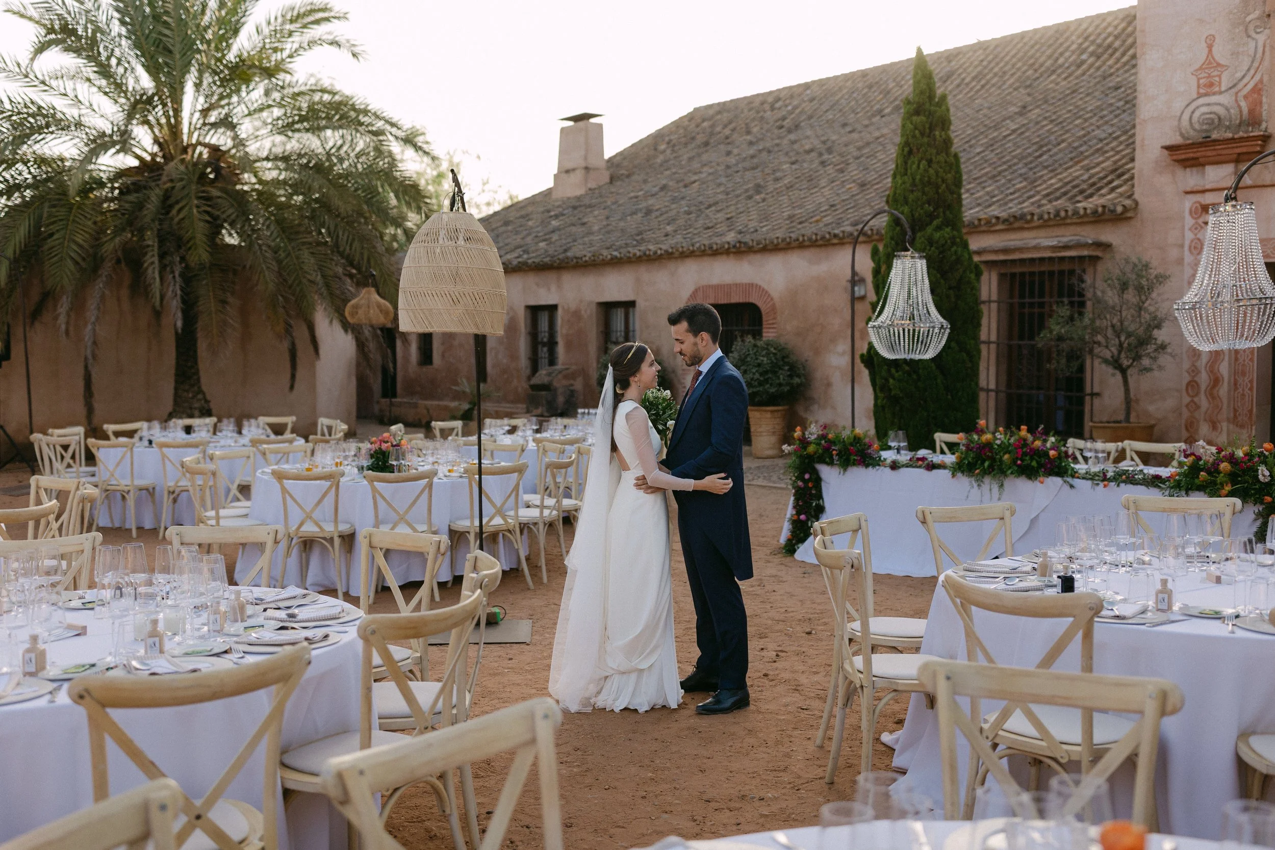 Pareja de recién casados en boda al aire libre, rodeados de mesas decoradas con manteles blancos y vajilla, en un ambiente rústico con palmeras y construcciones antiguas.
