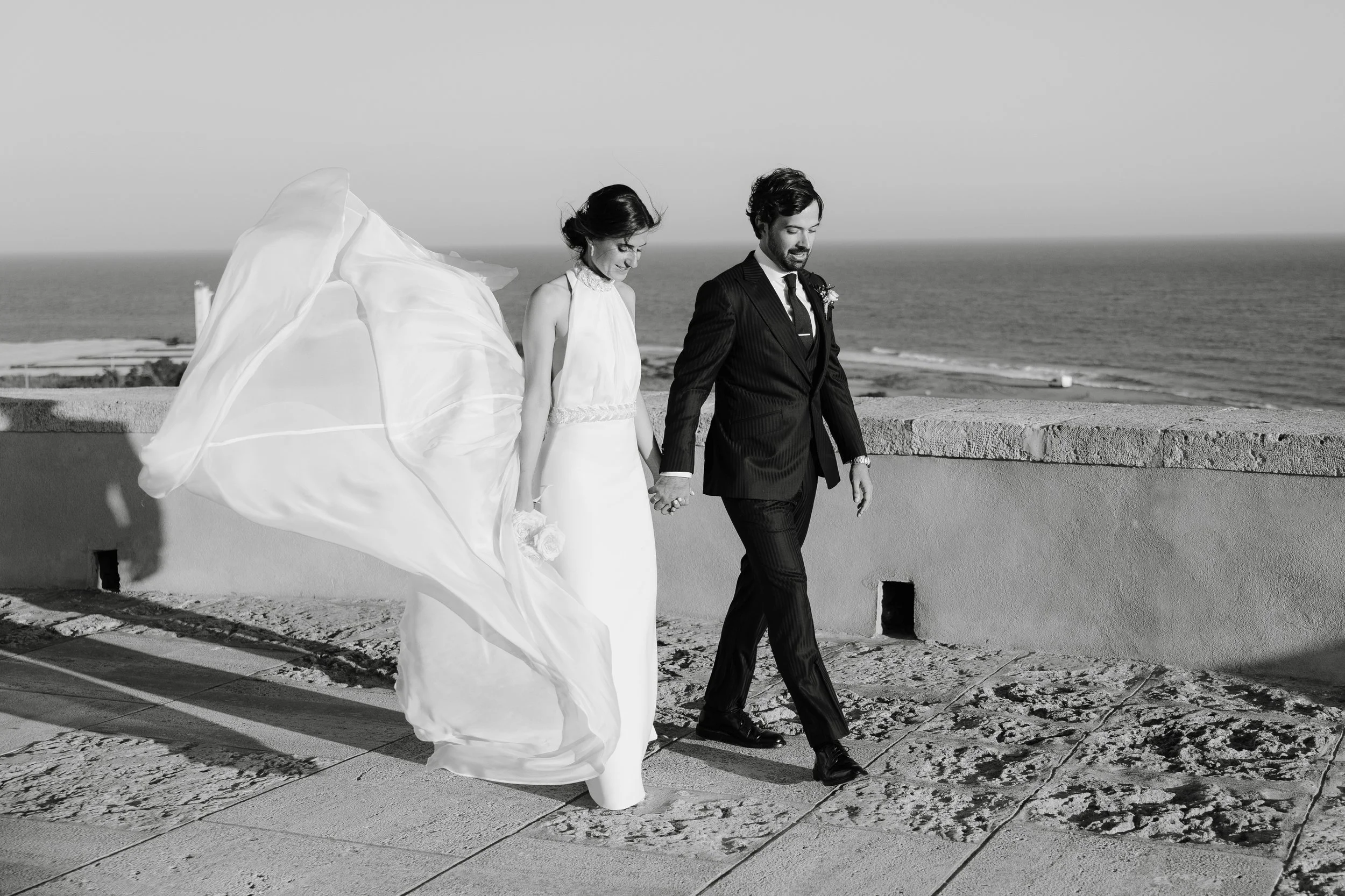 Una pareja de recién casados caminando por una terraza cerca del mar, en la playa, en una fotografía en blanco y negro.