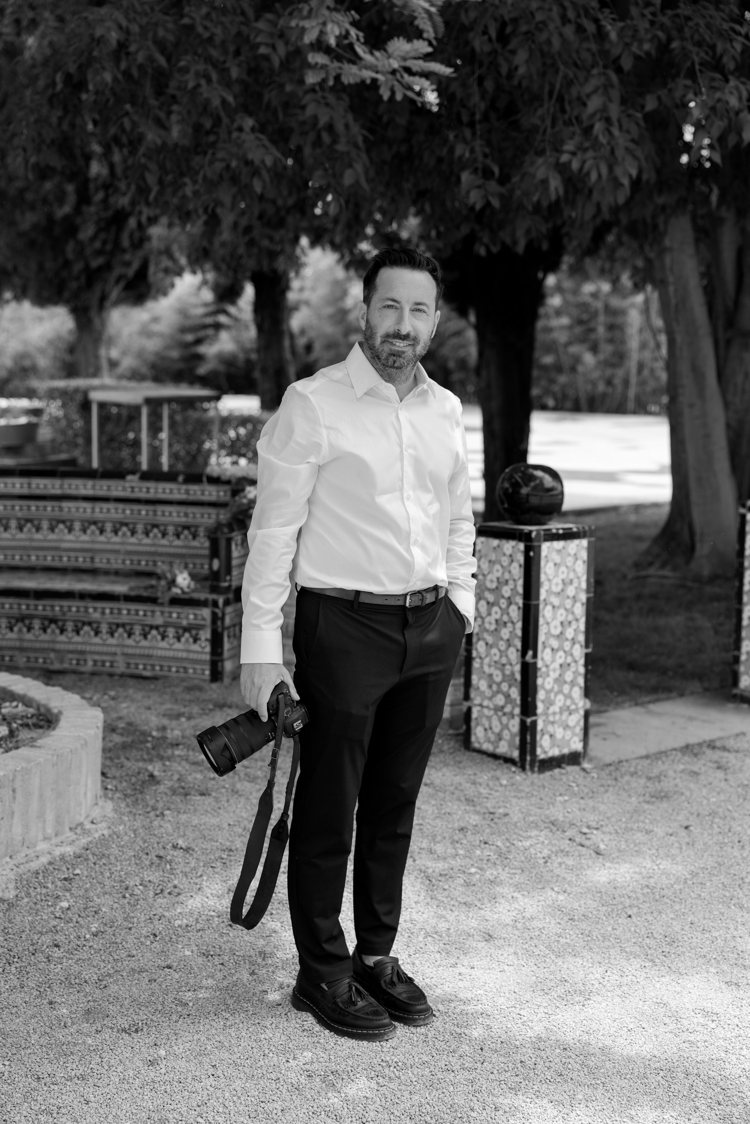 Un hombre con camisa blanca y pantalones oscuros sosteniendo una cámara en una mano, de pie en un entorno al aire libre con árboles y mobiliario decorativo, en blanco y negro.