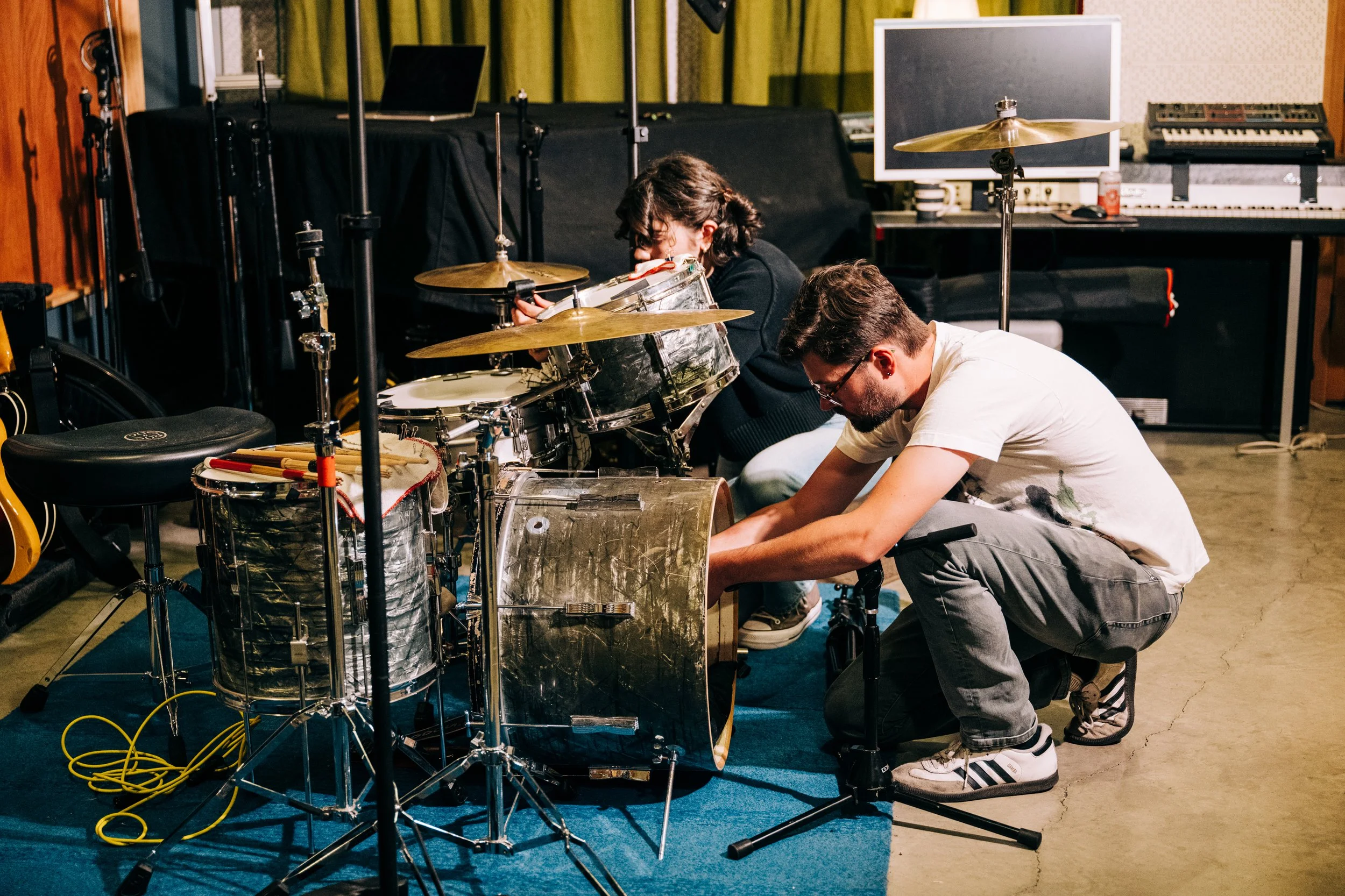 Two musicians working on a drum set in a music studio, with various instruments and equipment in the background.
