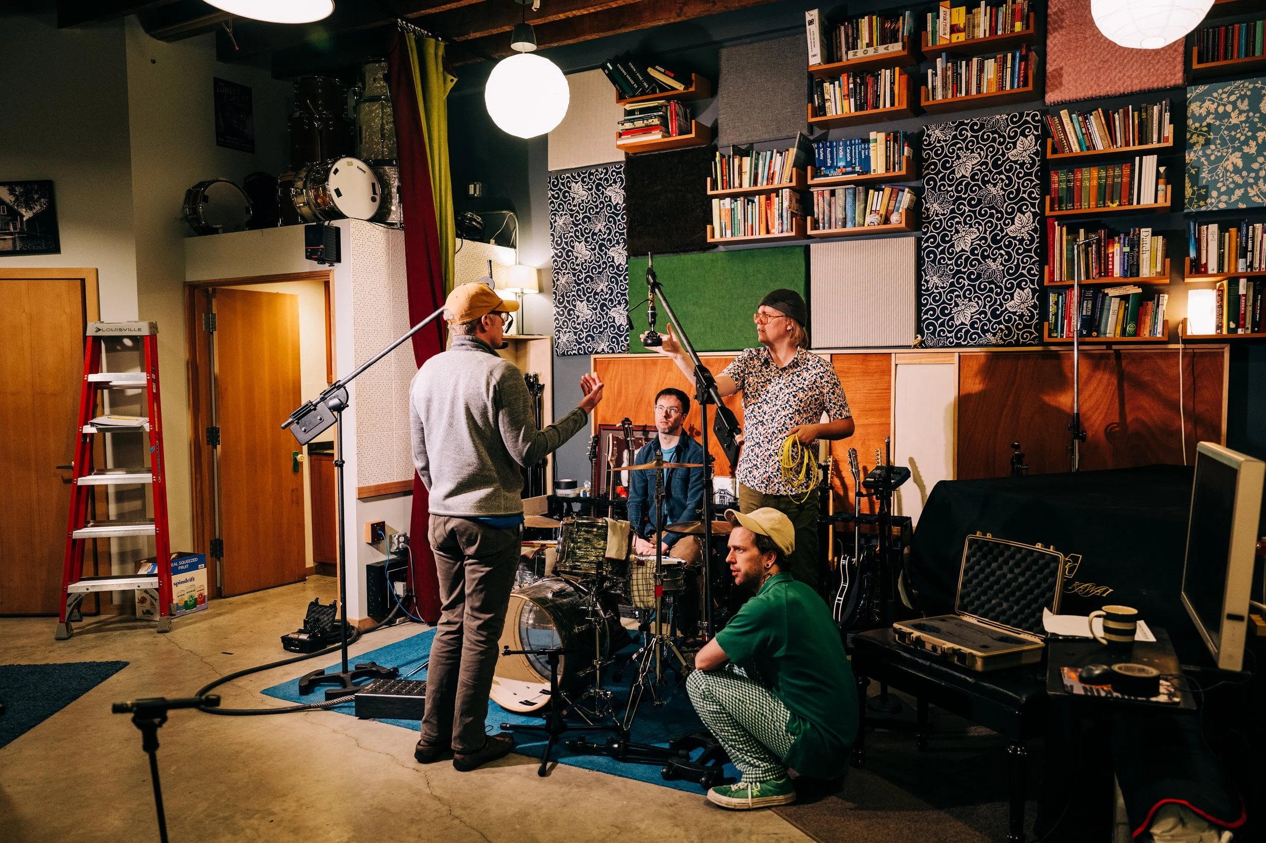 A music video or film crew is recording in a studio room with musical instruments, including drums, and equipment. Crew members are setting up and adjusting microphones and other gear, with a bookshelf filled with books and decorative panels on the w