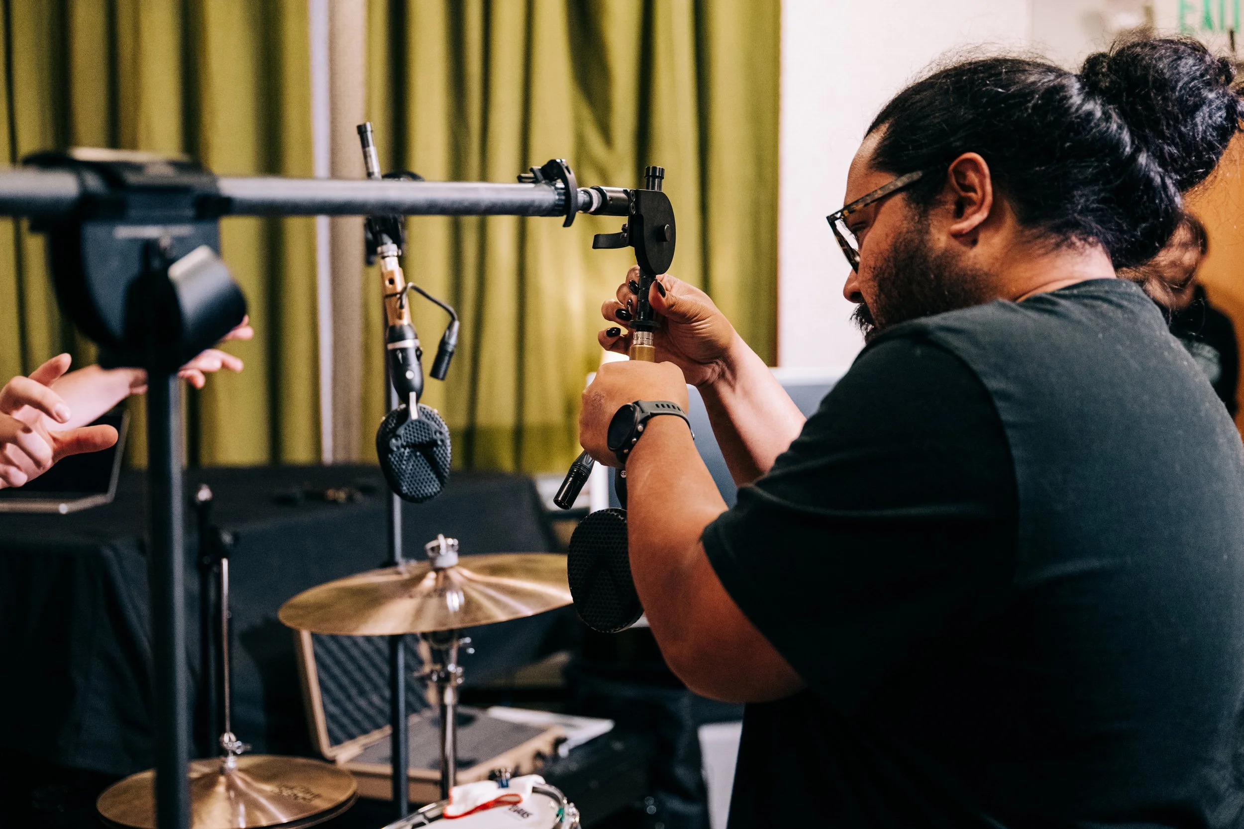 A man with glasses and a beard wearing a black shirt is adjusting a microphone stand. There are drums and a microphone on a stand nearby, with green curtains in the background.
