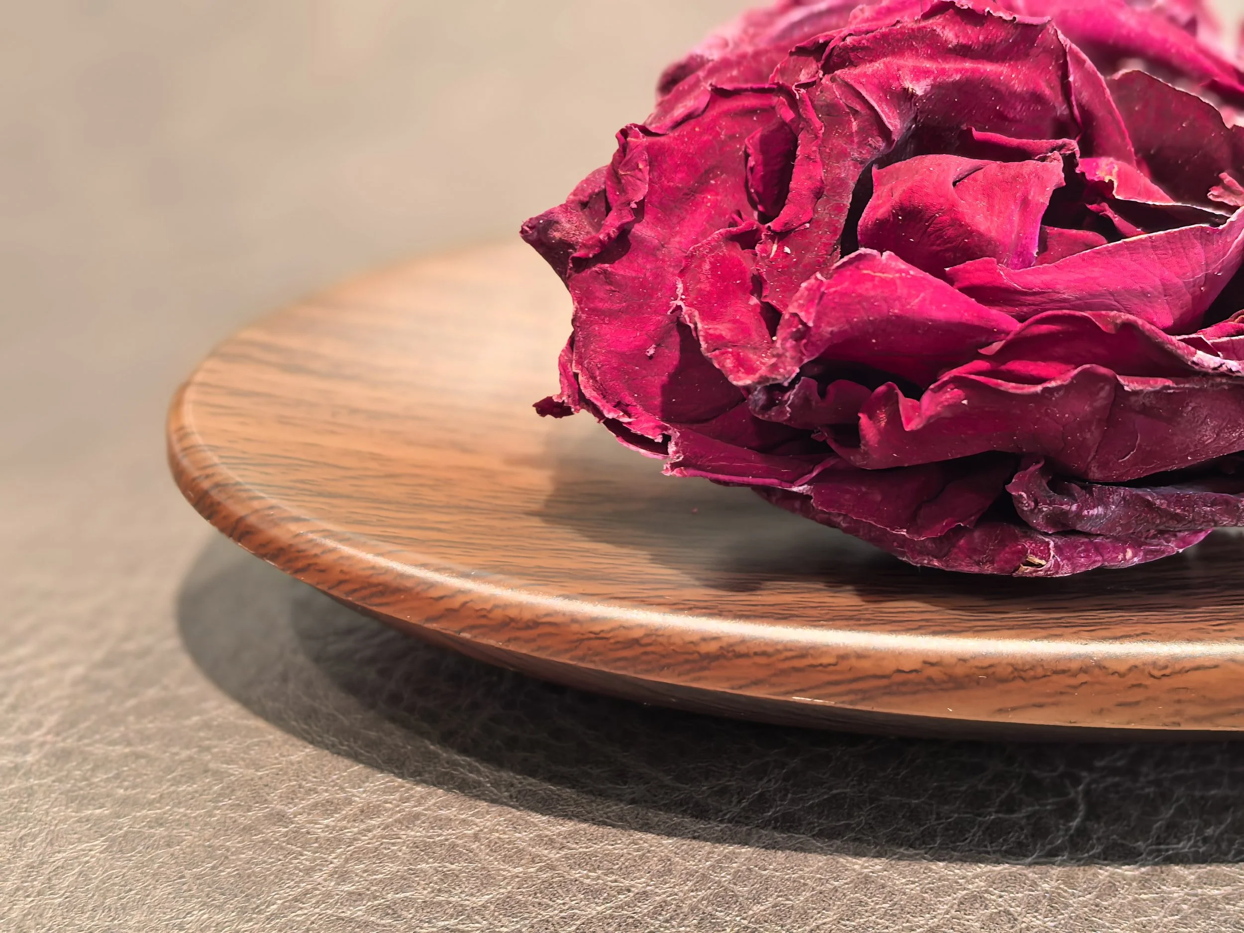 A close-up of a dried, dark pink rose on a wooden plate, with a plain beige background.