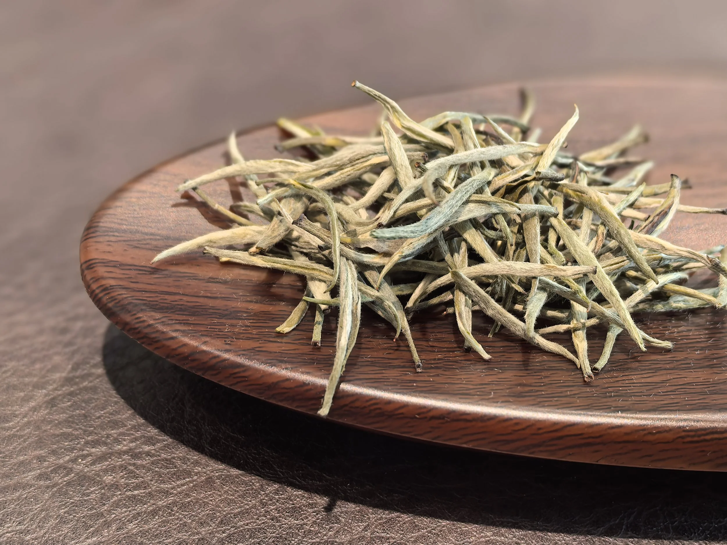 Dry, twisted, pale green tea leaves on a round wooden plate.