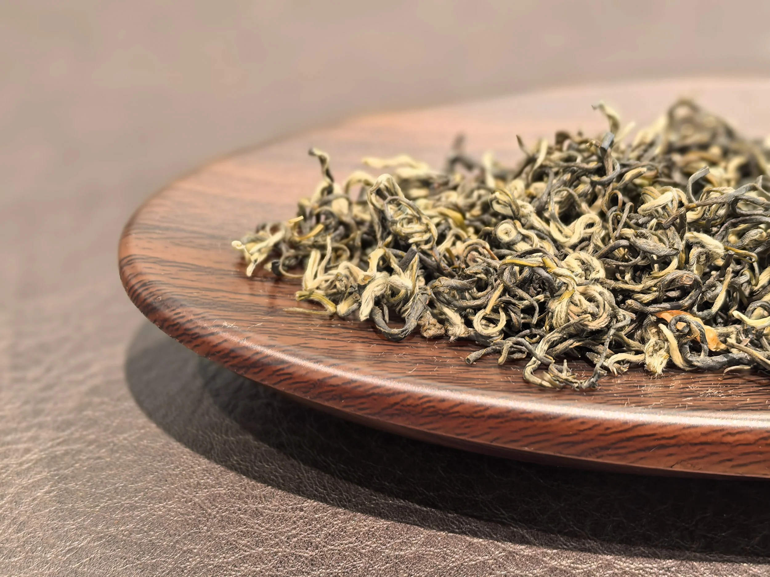 Dried leaves or herbal tea leaves spread on a round wooden plate, placed on a textured brown surface.