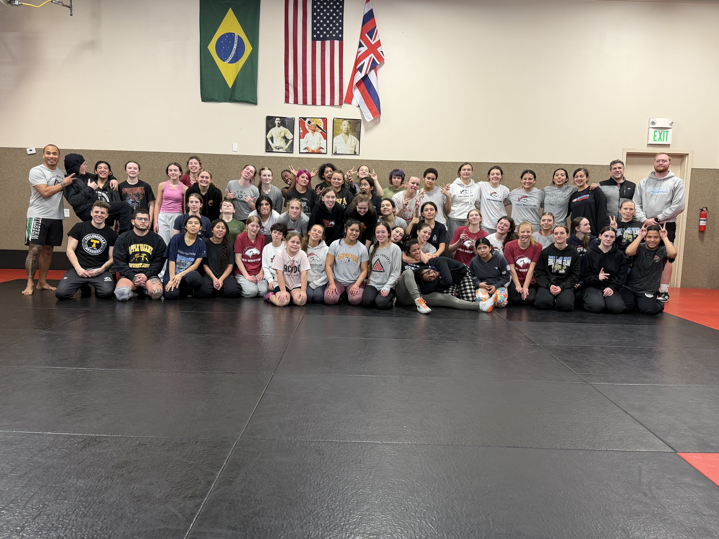 Group of young women and men in a martial arts gym, some in workout attire, posing for a group photo on black mat flooring with flags of Brazil, the United States, and the United Kingdom hanging on the wall behind them.