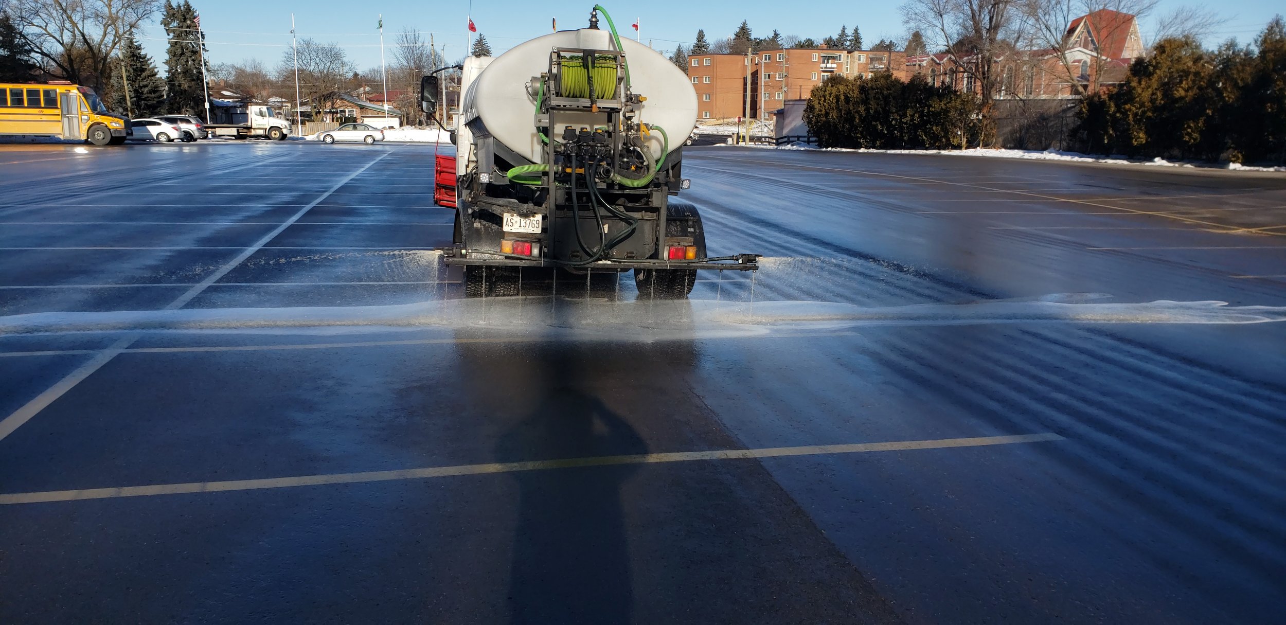A parking lot being salted or de-iced by a street maintenance vehicle with a large tank and spraying equipment, with snow and ice on the ground.