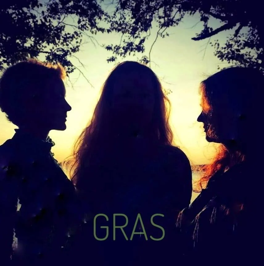 Silhuetter av tre kvinner mot solnedgang med trær i bakgrunnen, med teksten 'GRAS' nederst.