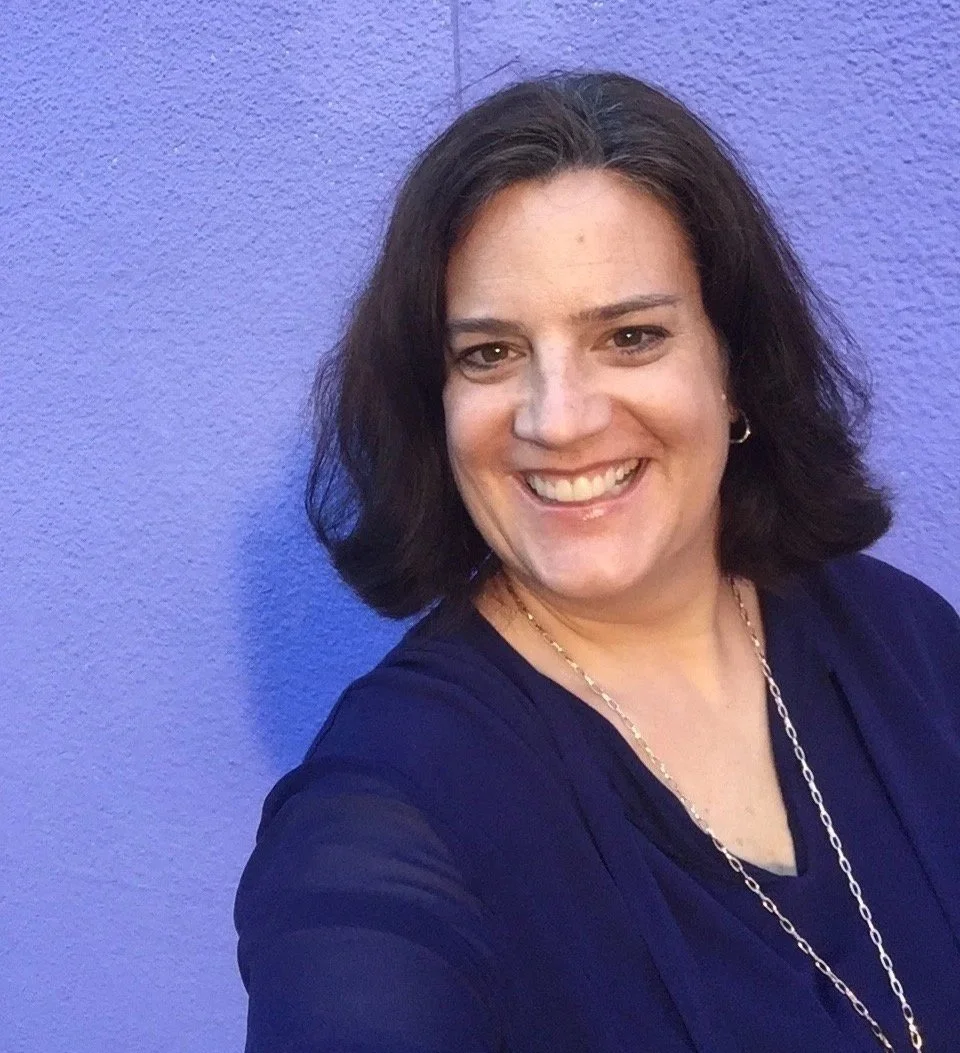 A smiling woman home organizer in Denver and Boulder who loves to help people declutter, organize and thrive with shoulder-length dark hair, wearing a dark top and a gold chain necklace, standing against a textured purple wall.