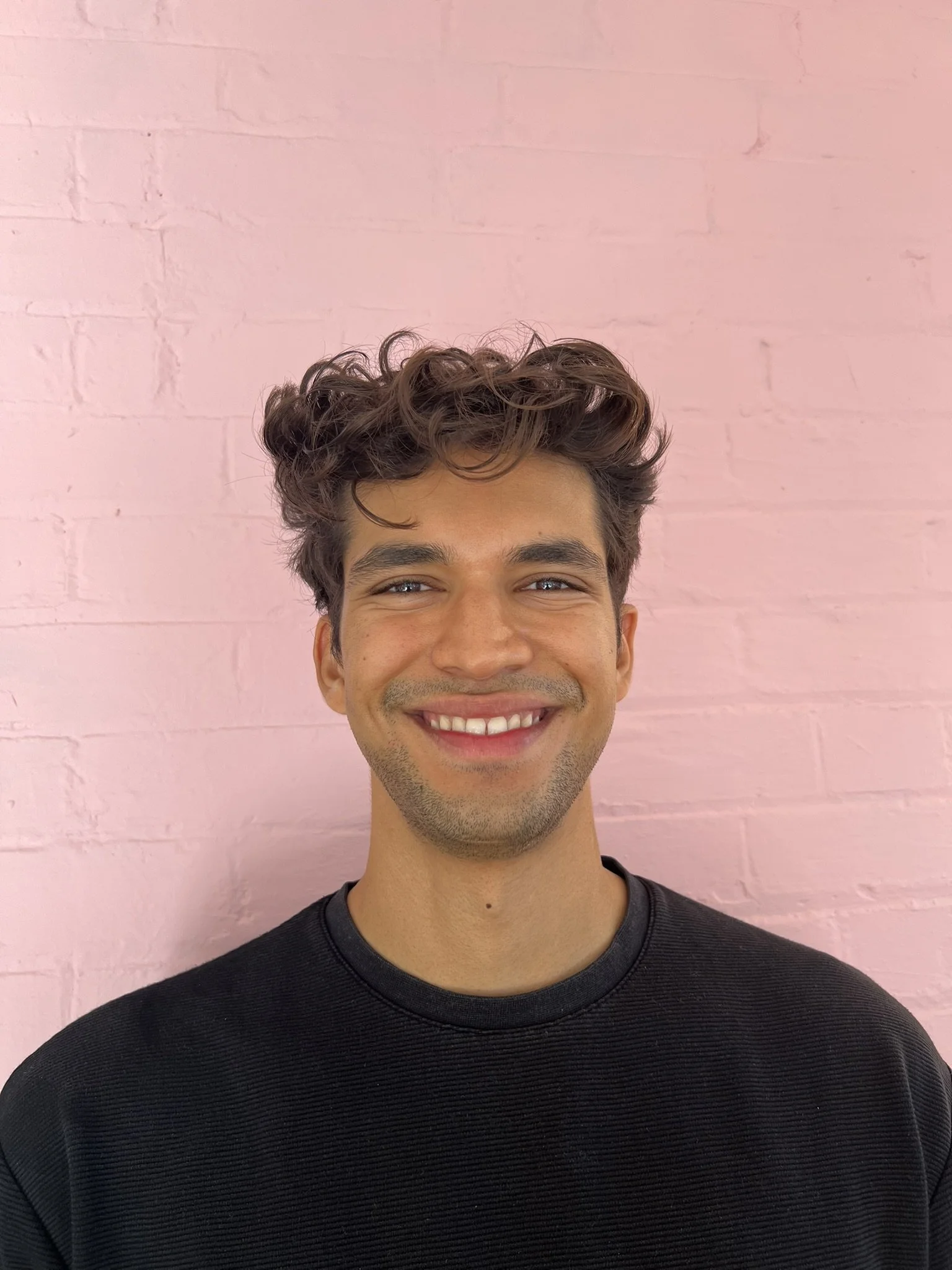 A young man with curly brown hair smiling in front of a pink brick wall.