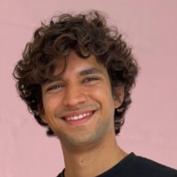 Young man with curly hair smiling against a pink background.