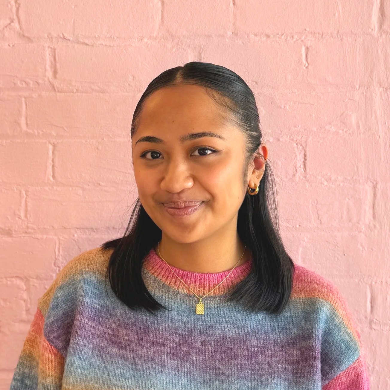 Young woman with shoulder-length dark hair, wearing gold hoop earrings and a gold necklace with a rectangular pendant, smiling in front of a pink brick wall, dressed in a multicolored rainbow striped sweater.