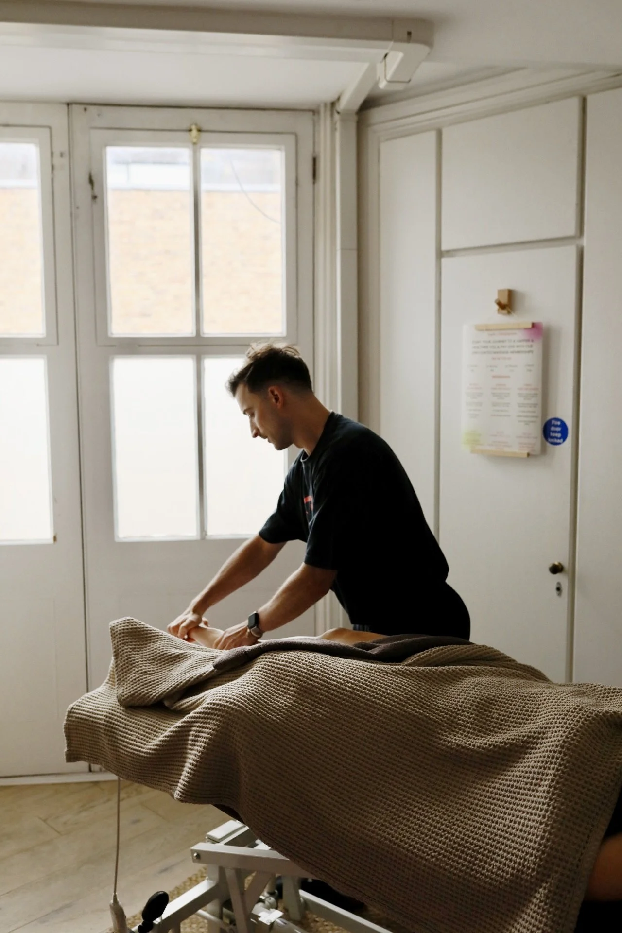 A person in black scrubs making a bed in a room with a large window.