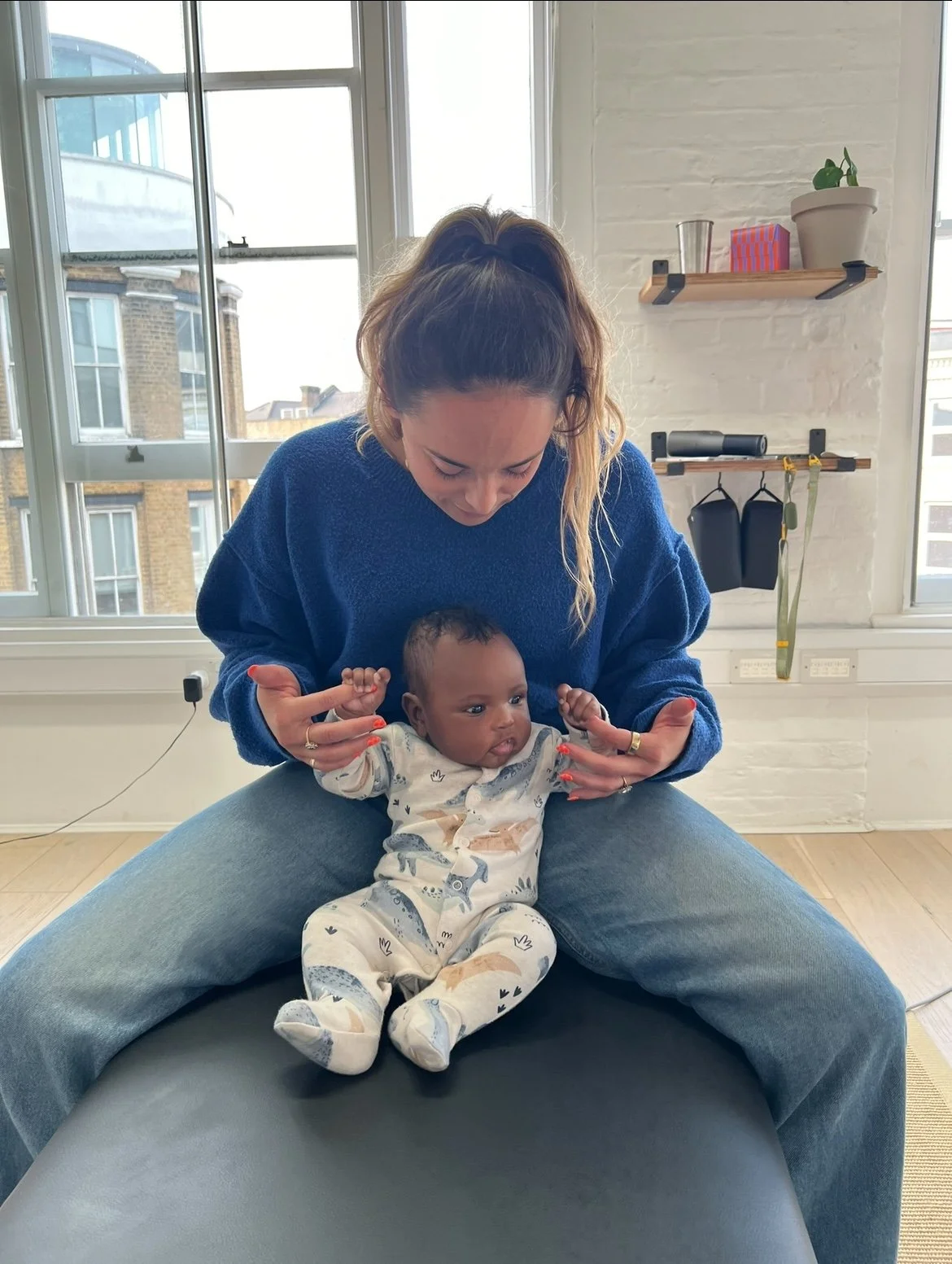 A woman sitting on a large round cushion, holding a baby on her lap inside a bright room with large windows and a view of neighboring buildings. The woman wears a blue sweater and has her hair tied back, while the baby is dressed in a patterned onesie.