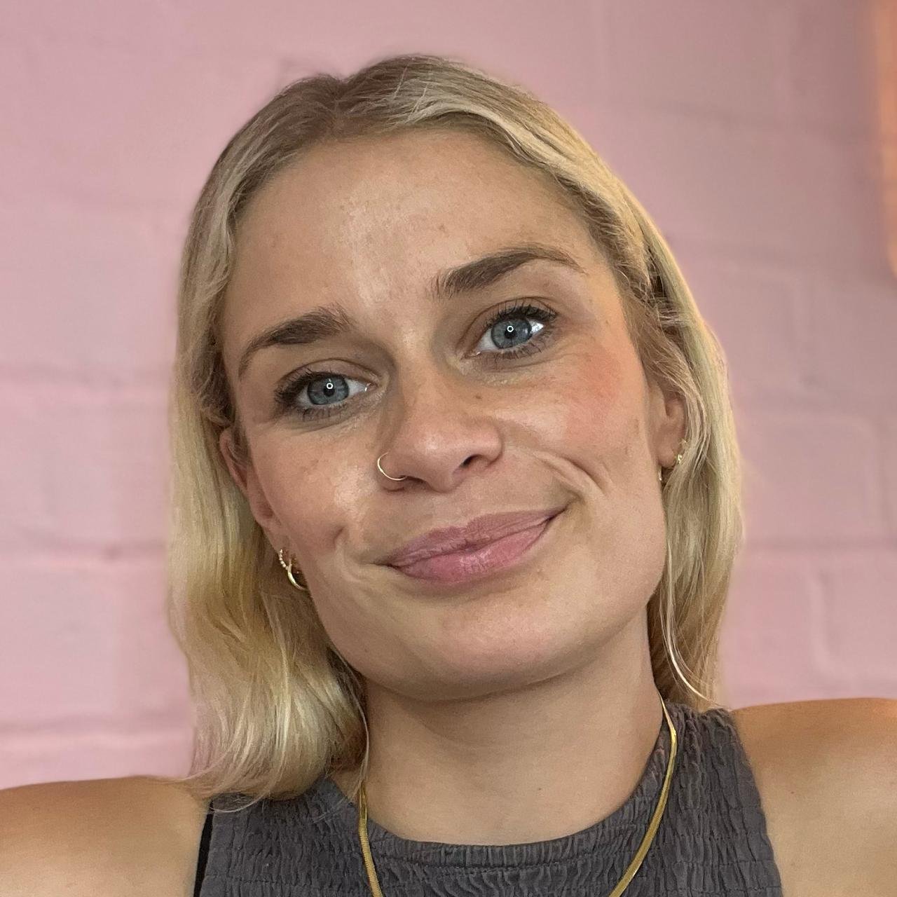 A young woman with blonde hair and blue eyes smiling at the camera, wearing small hoop earrings, a nose ring, and a black sleeveless top, against a pink background.