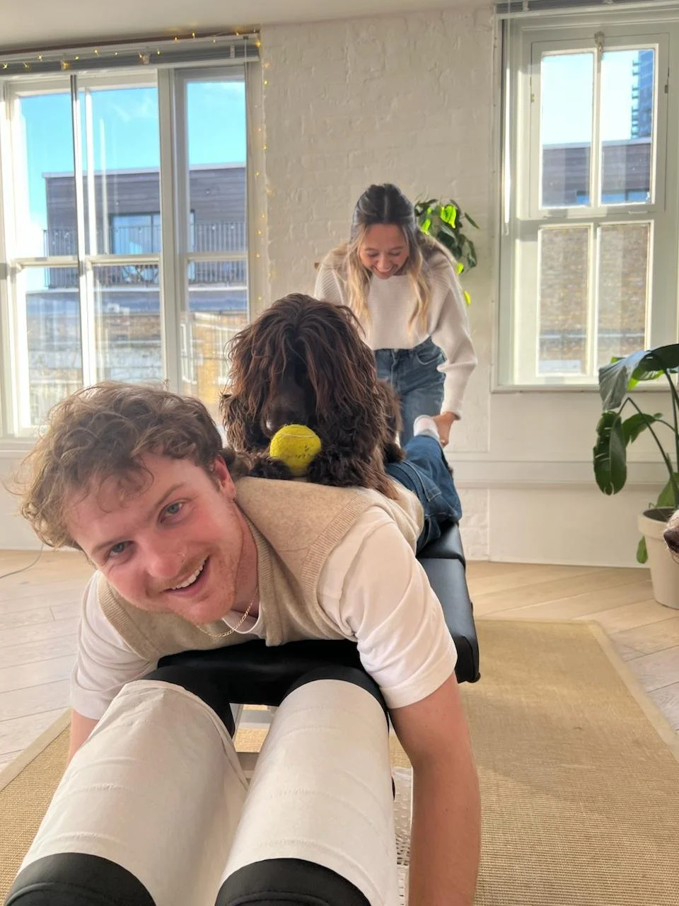 A man lying face down on a massage table with a dog lying on his back. The dog has a yellow ball in its mouth. Two women are smiling and interacting with the dog in a bright, cozy room with large windows and potted plants.