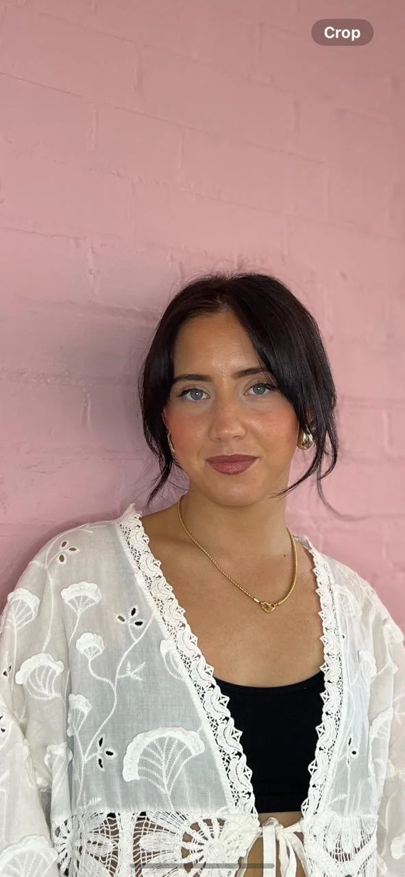 A woman with dark hair and blue eyes, wearing a white embroidered jacket over a black top, standing against a pink brick wall.