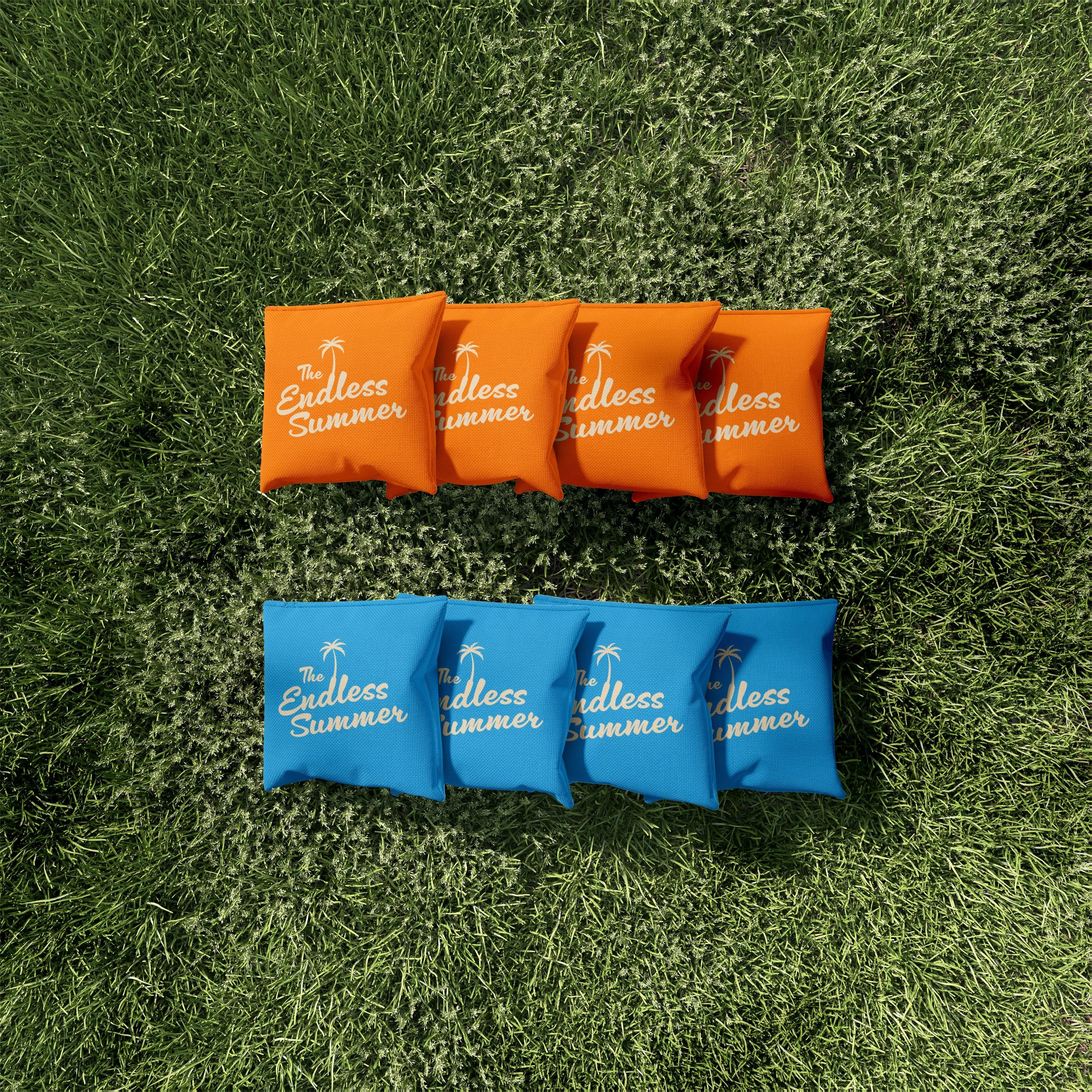 Four orange beach towels with white text and a palm tree logo, laid out on green grass. The towels read "The Endless Summer."