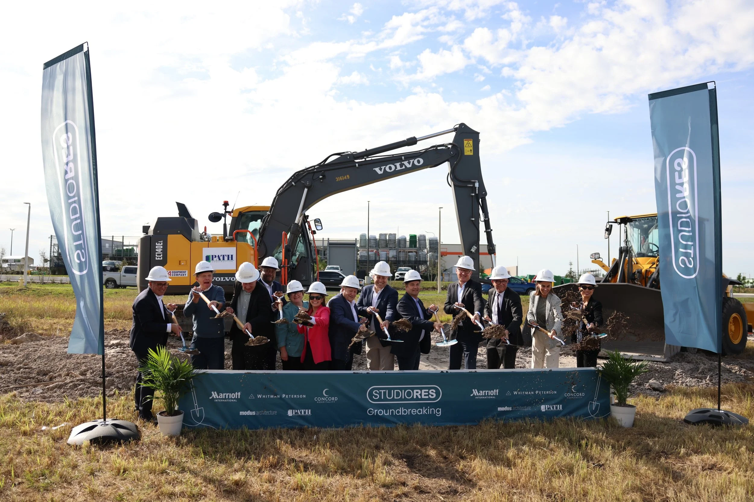 A group of people in formal attire and safety helmets participating in a groundbreaking ceremony for a construction site, with excavation equipment and flags displaying the event branding.
