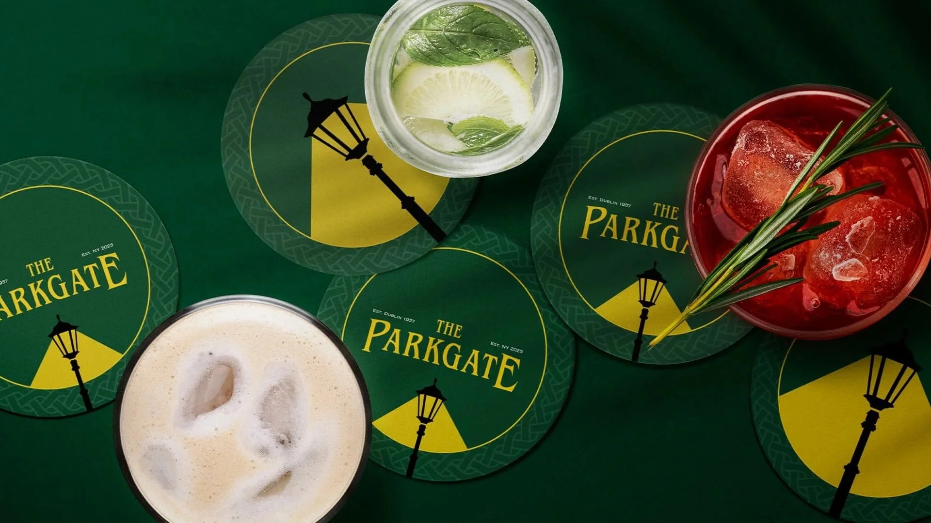 Top-down view of two glasses of cocktails with ice, one with lemon and mint, the other with large ice cubes and garnished with a sprig of rosemary, surrounded by round coasters displaying a logo for The Parkgate with a lamp post graphic.