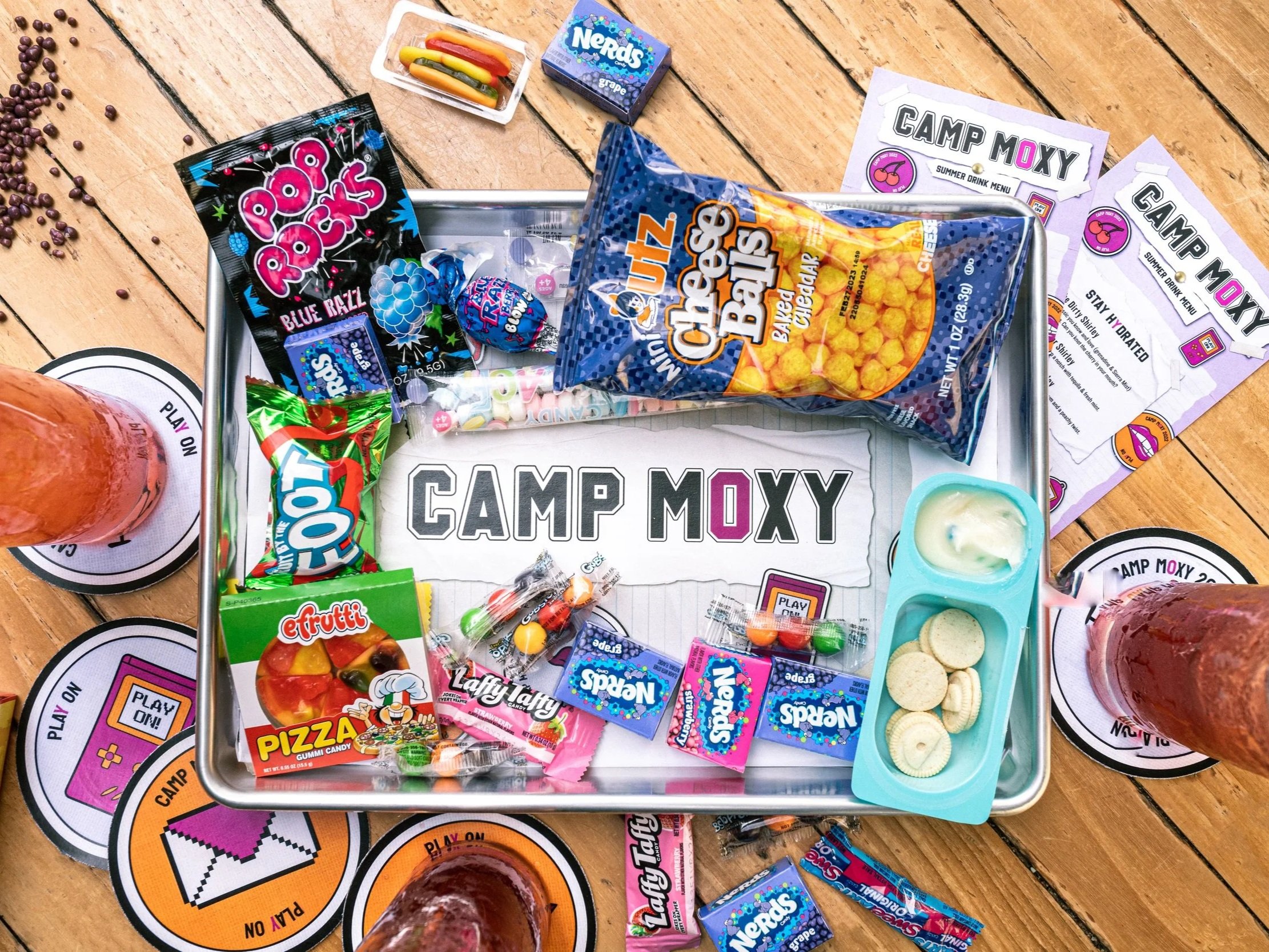 A table set for a camping-themed snack, with a metal tray holding candy, snacks, and a paper towel with 'CAMP MOXY' printed on it. Around the tray are cups with ice and drinks, and circles with 'PLAY ON' and 'CAMP MOXY' logos. Items include gummy can