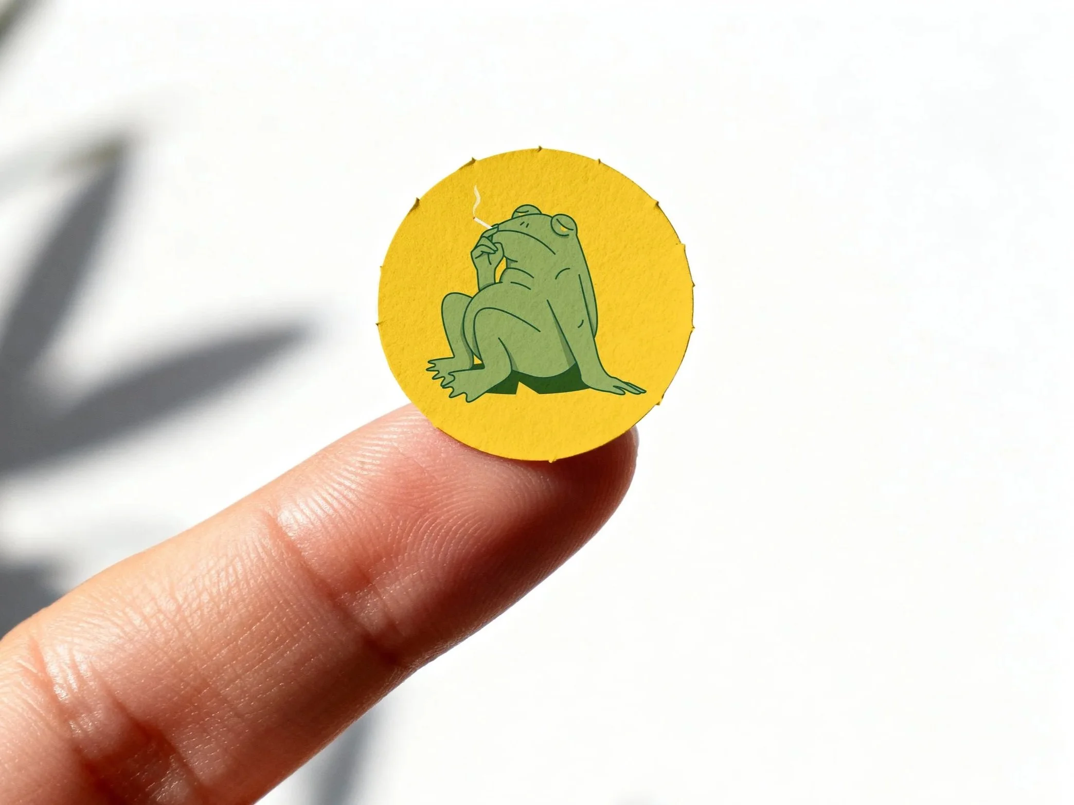 A hand holding a circular yellow object with a cartoon illustration of a frog smoking a cigarette