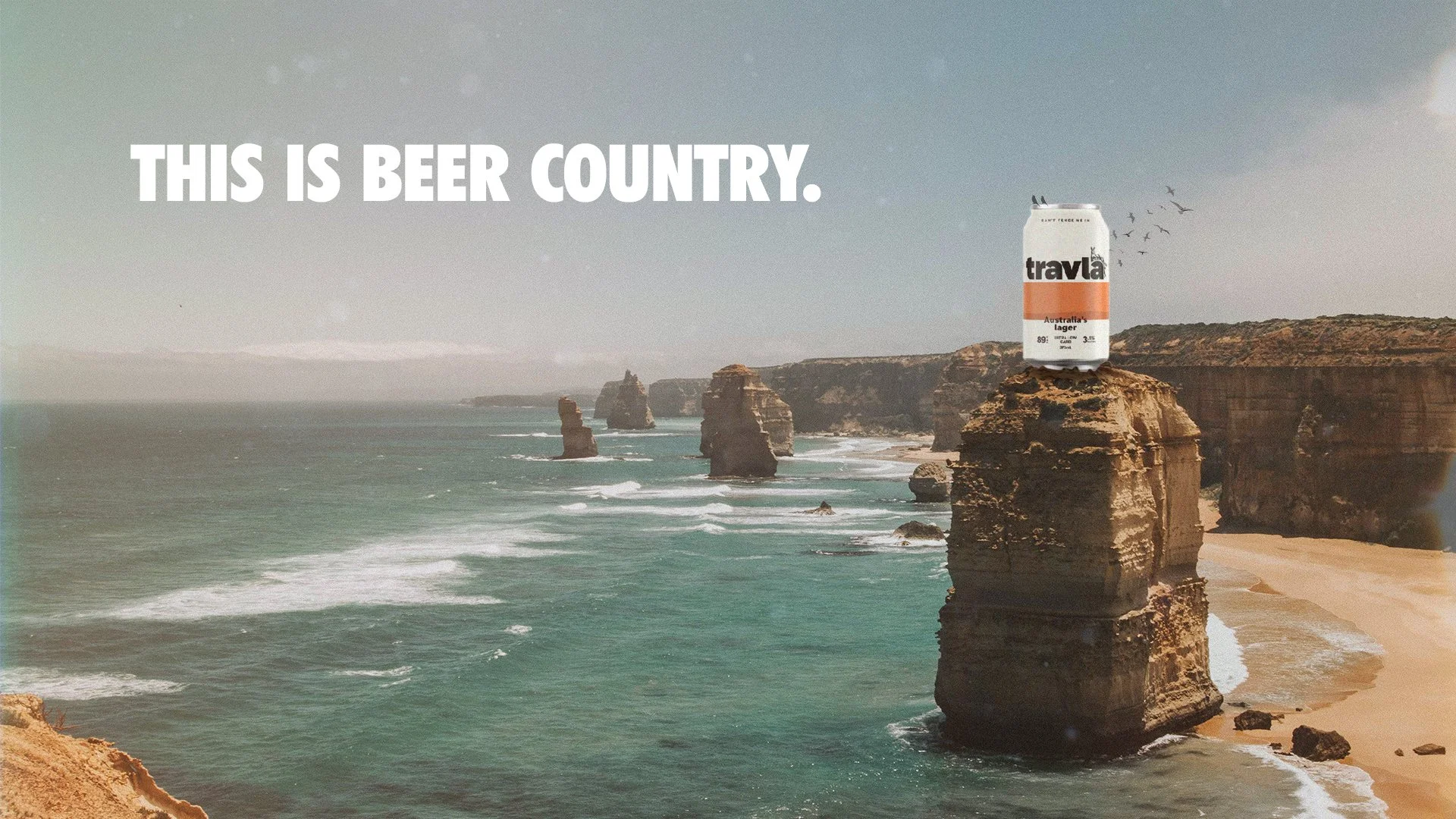 A large can of Travla Australia’s Lager beer on top of a rock formation on the Great Ocean Road, with the ocean, cliffs, and rock formations in the background, and the phrase "This is Beer Country."