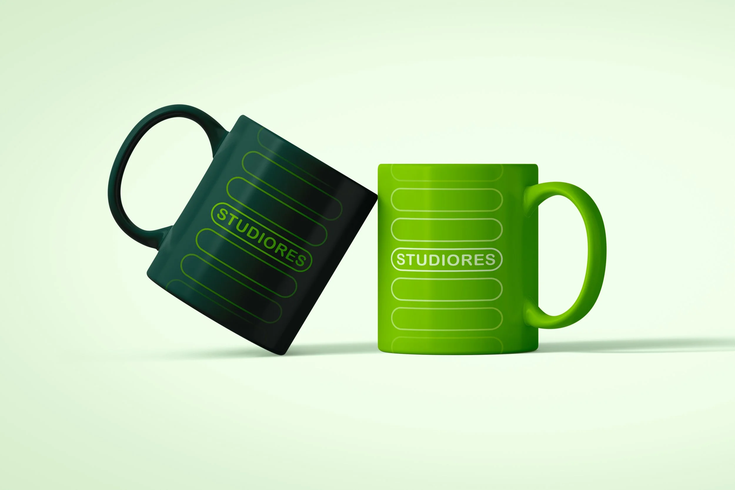 Two coffee mugs, one black and one green, with the word 'STUDIORES' printed on them and designs of horizontal lines. The mugs are tilted on a light background.