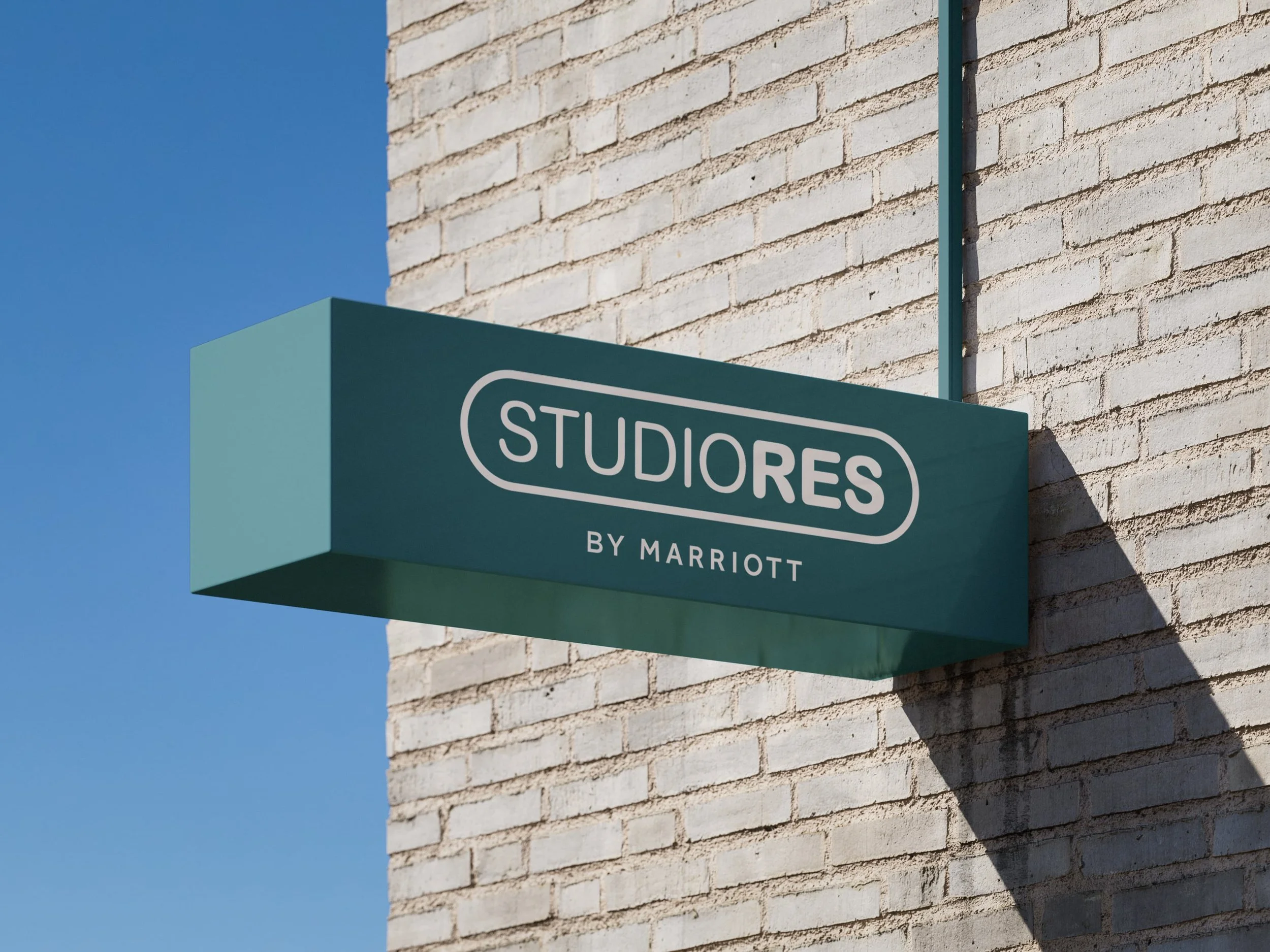 Close-up of a teal-colored sign with white text reading 'STUDiORES' attached to a brick wall, with part of the text 'BY MARRIOTT' visible at the bottom. The sky is clear and blue in the background.