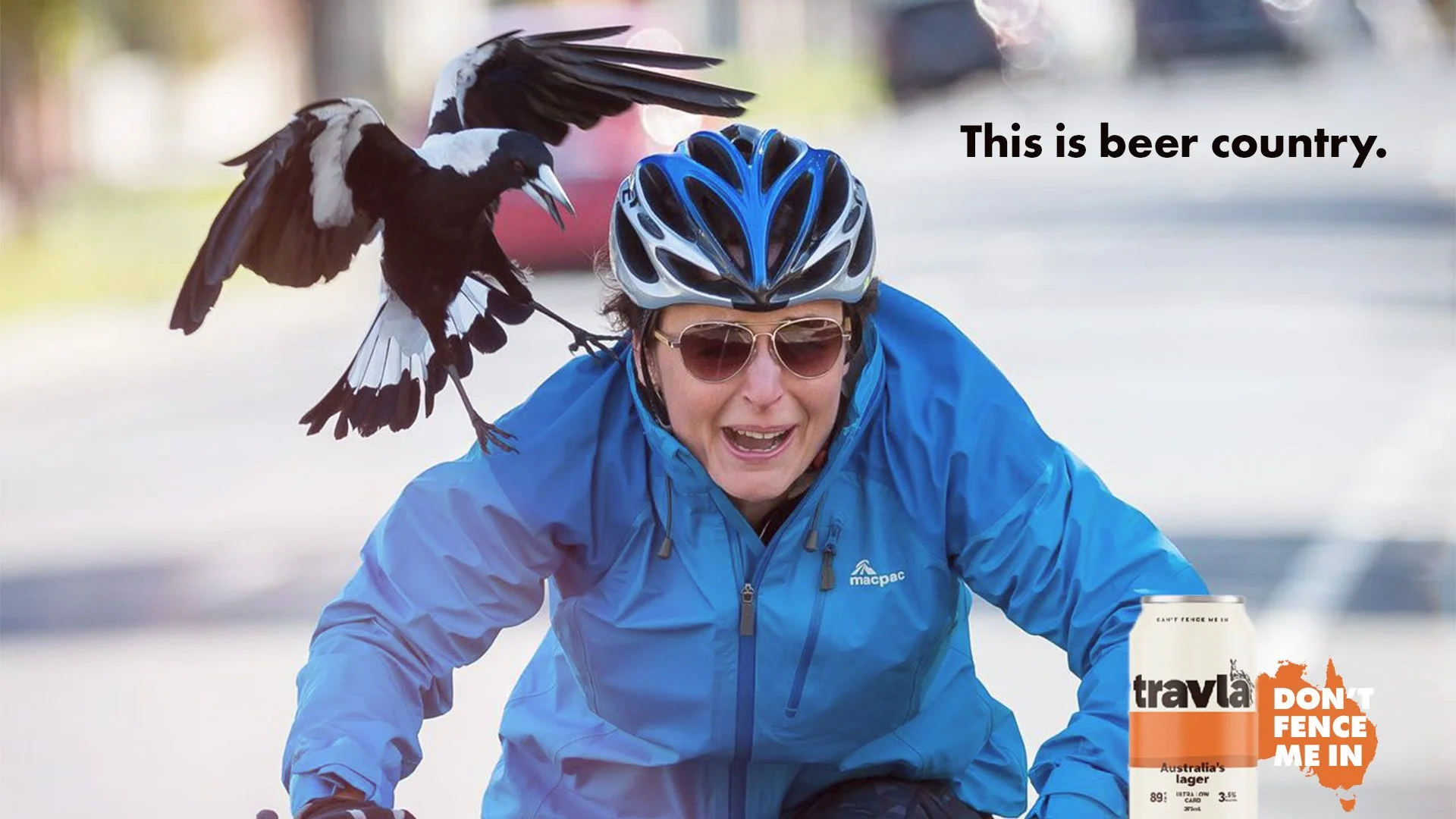 A woman wearing a blue jacket, sunglasses, and a bicycle helmet, smiling while riding a bike. A magpie with black and white plumage is perched on her shoulder. In the background, there is a can of Tralya lager and a sign that says 'Don't Fence Me In,