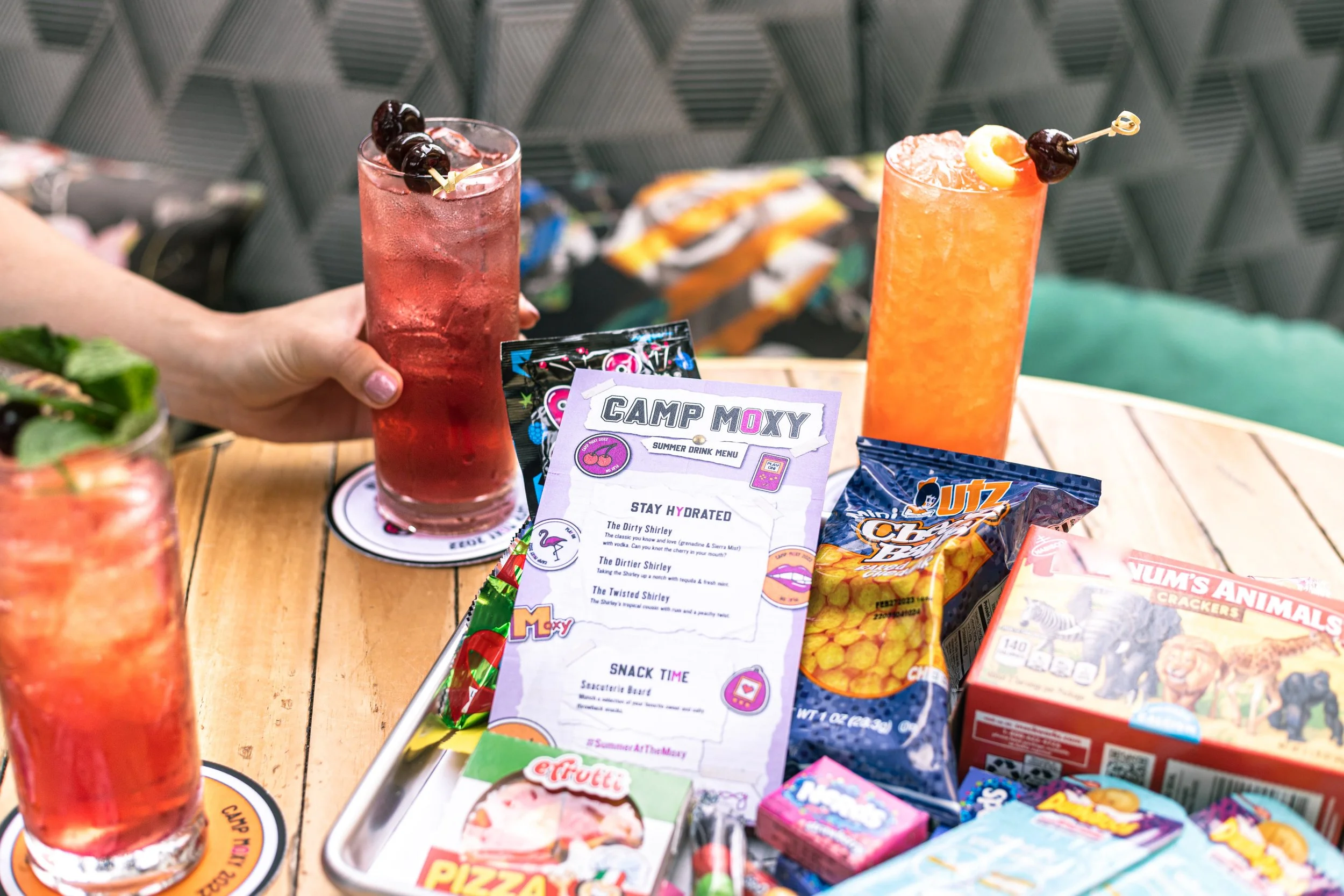 A hand holding a tall glass of red-colored cocktail with ice and three dark cherries on a skewer. Several other colorful drinks and snacks are on a wooden table, including chips, crackers, and a snack box, with a map of some sort and a colorful summe