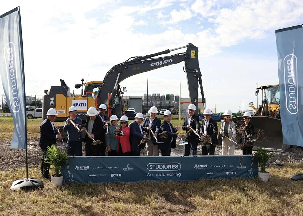 Group of people in formal attire and white safety helmets participating in a groundbreaking ceremony with shovels, in front of construction equipment and flags.
