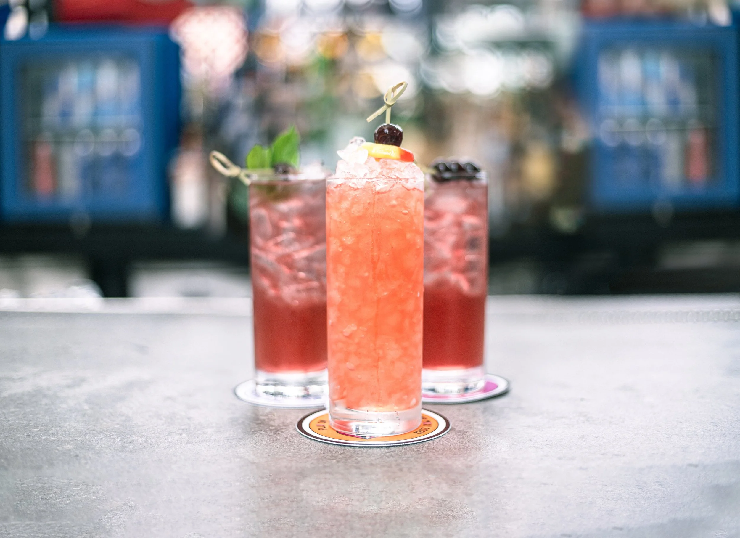Three tall glasses of pink cocktails with ice, garnished with berries, mint, and citrus slices, on a gray table outdoors with a colorful blurred background.
