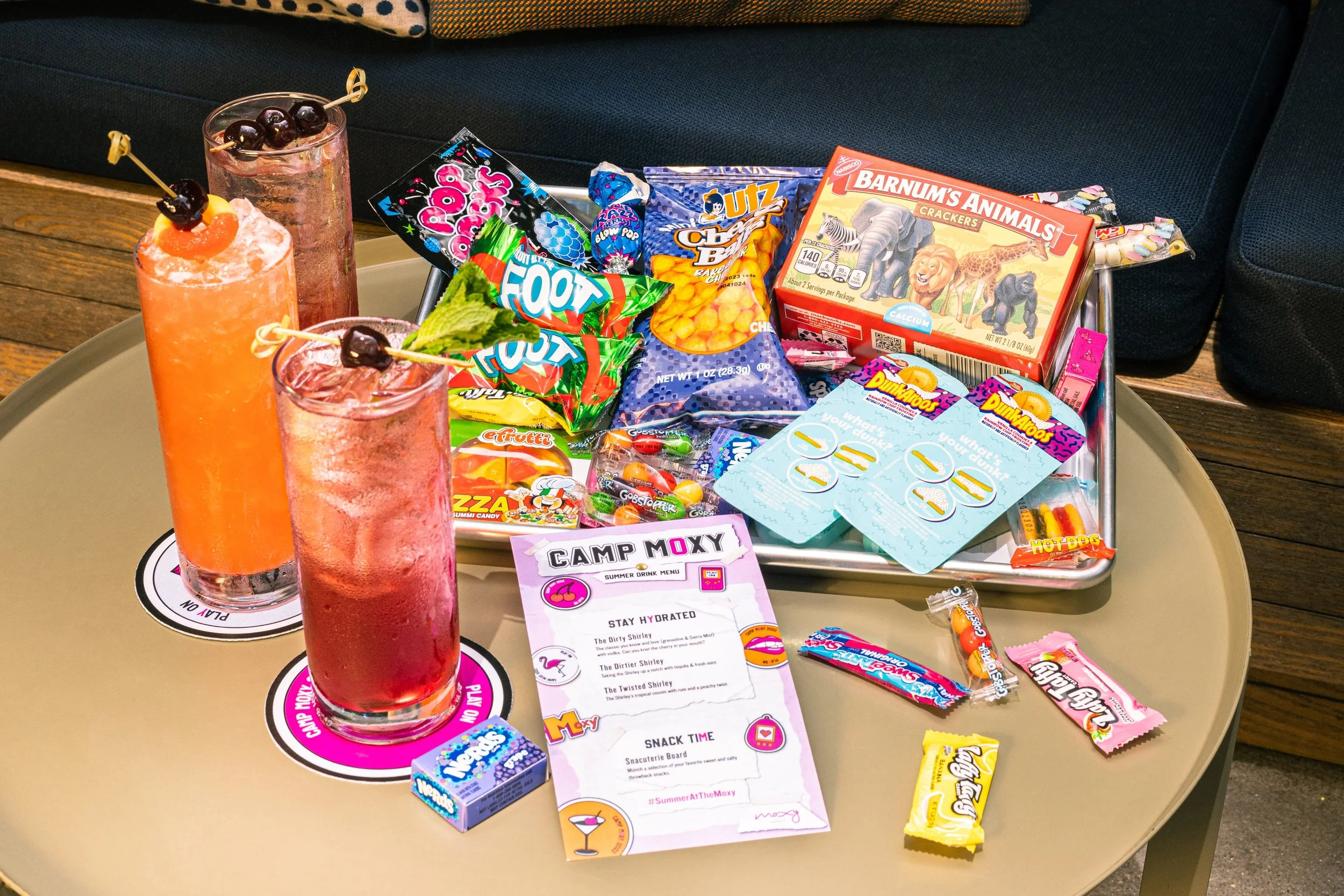 Tray with various snacks including candy, chips, and a box of animal crackers, with three colorful drinks on a table.