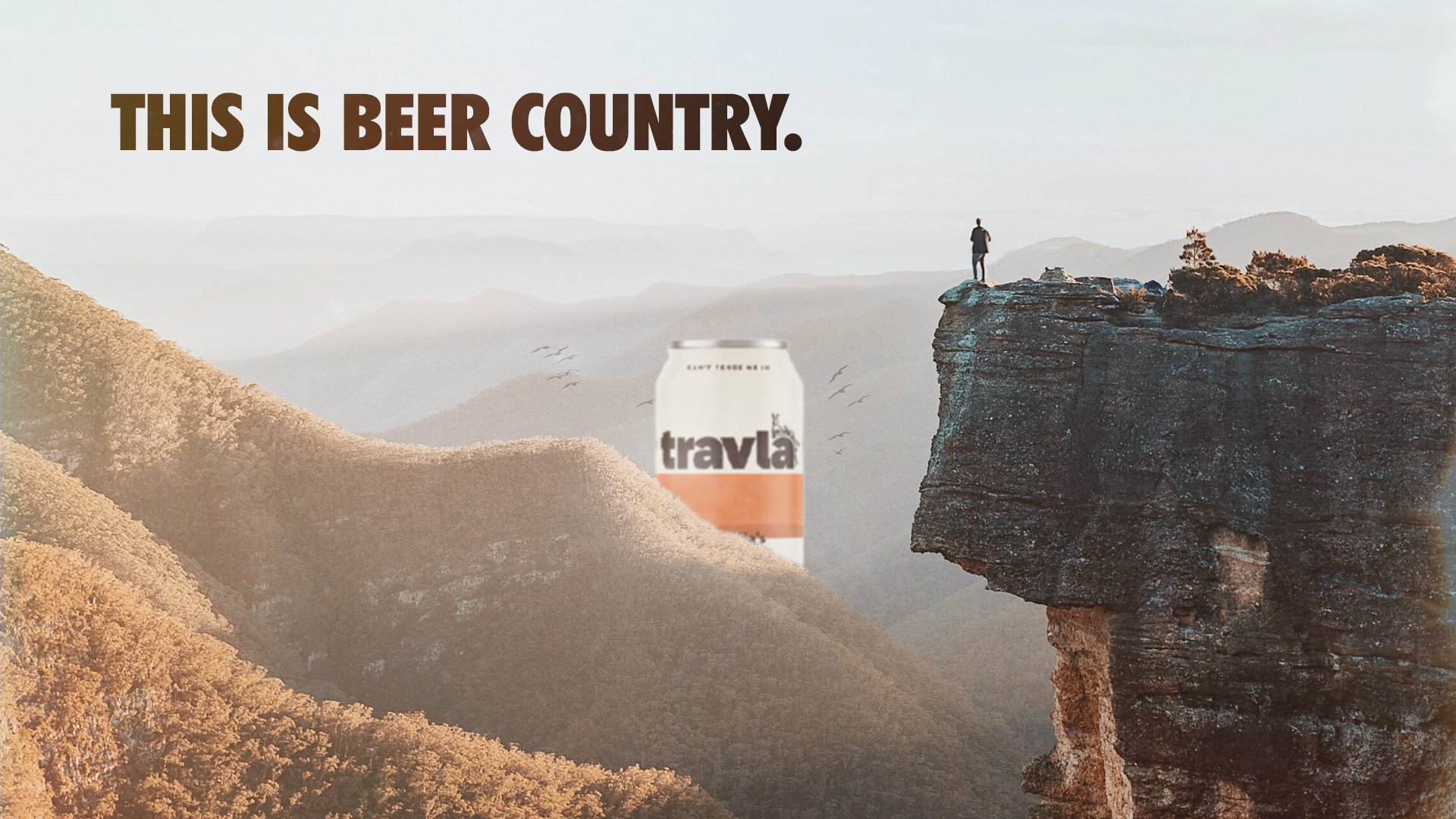 A hiker standing on a cliff overlooking a mountain landscape with layers of mountains in the distance. A can of Travla beer is edited into the scene, and a bold text in the upper left reads 'THIS IS BEER COUNTRY.'